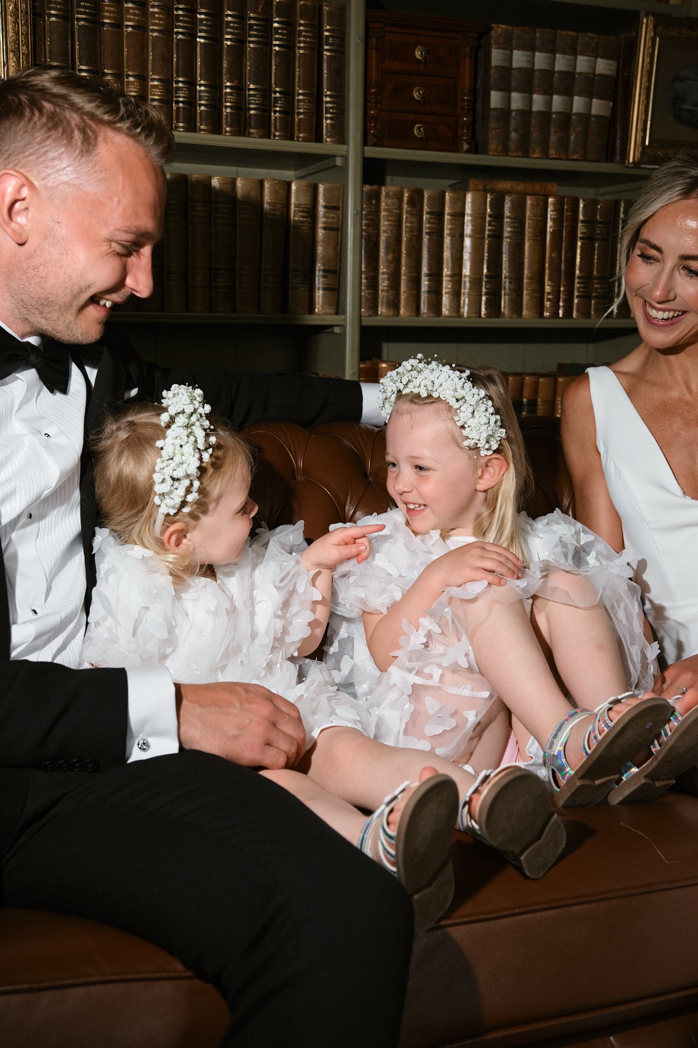 Familj i formella kläder, två vuxna och två unga flickor, sitter på en läderfåtölj i ett bibliotek. Flickorna bär vita klänningar och blomsterkransar, vuxna i formella kläder, alla ler och pratar.
