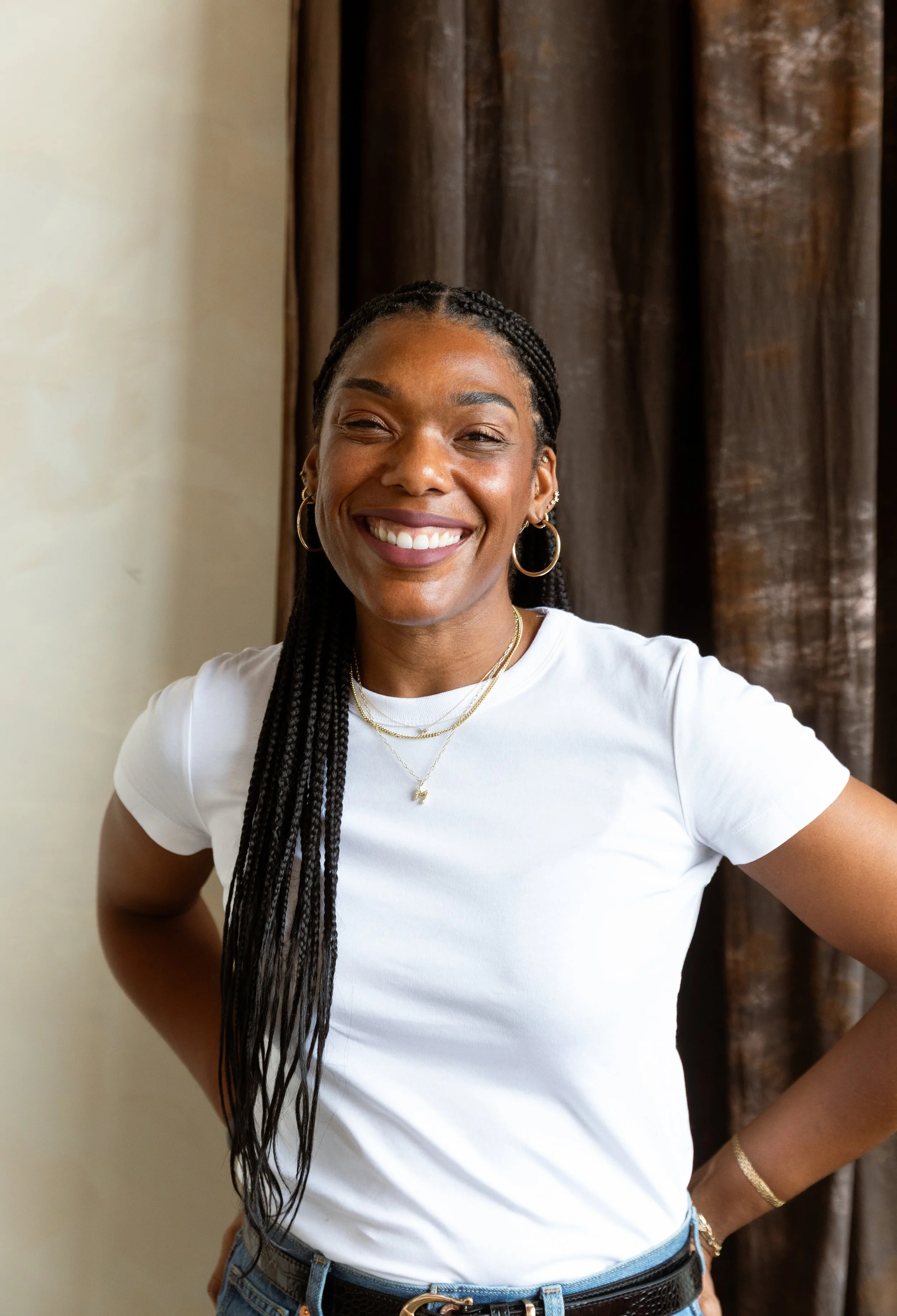 A woman with dark braided hair, wearing a white t-shirt, gold hoop earrings, layered necklaces, and a gold bracelet, smiling and standing indoors in front of a beige wall and a brown curtain.