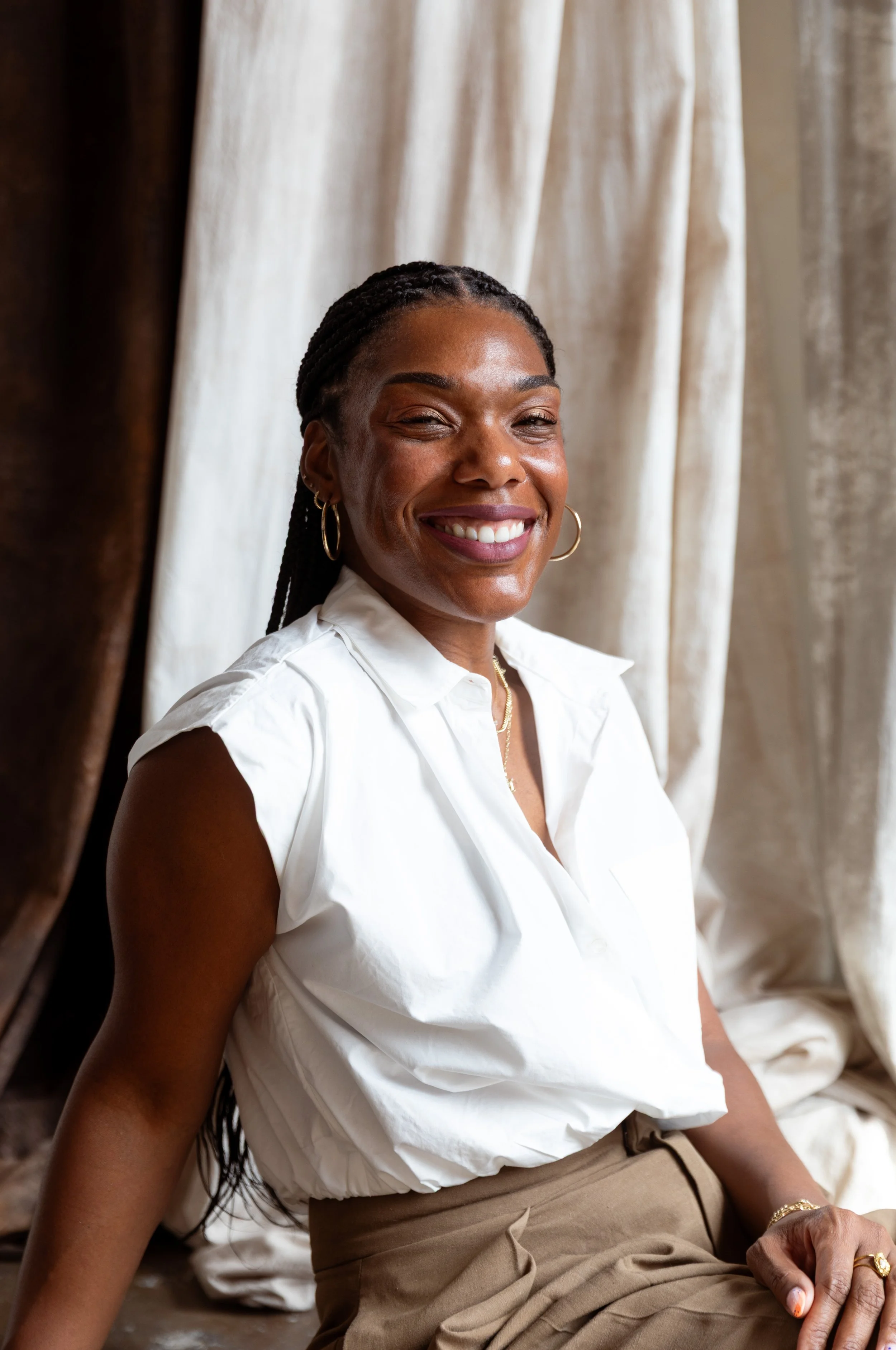 A smiling woman with dark braided hair, wearing gold hoop earrings and a white sleeveless blouse, sitting in front of beige and white curtains.