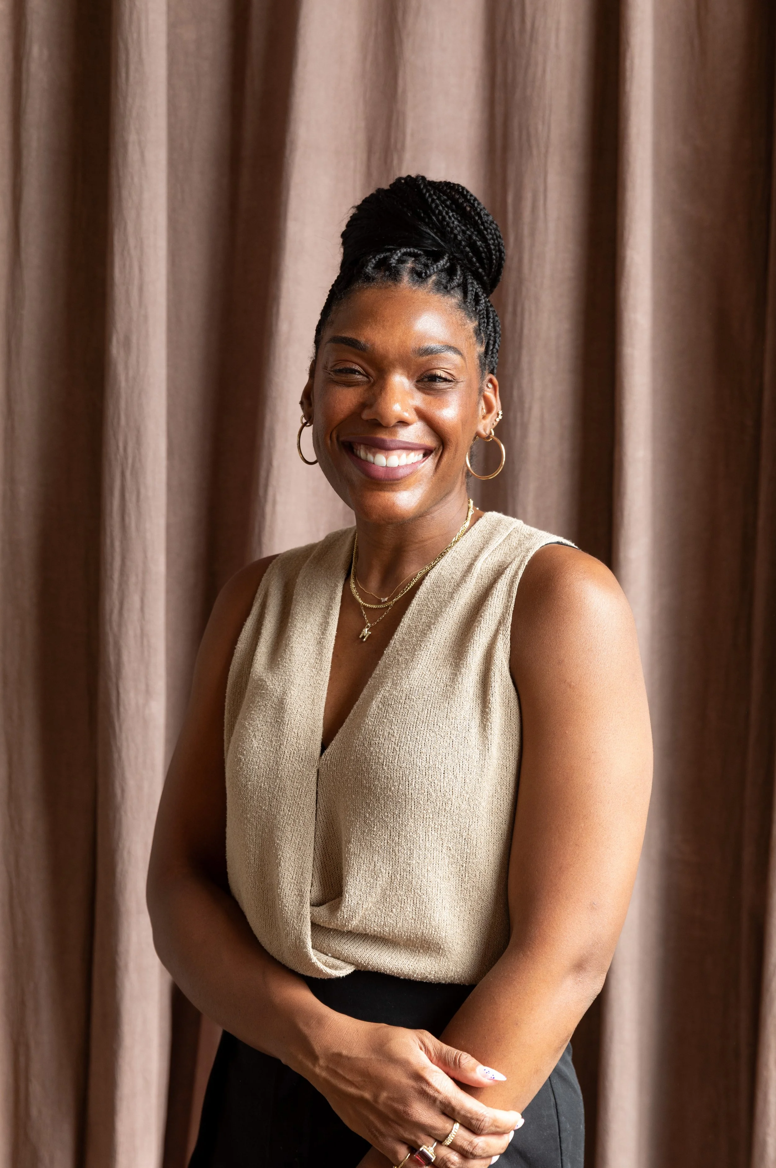 A woman with braided hair styled in a high bun, smiling, wearing hoop earrings, layered necklaces, a sleeveless beige top, and black pants, standing in front of a brown curtain.