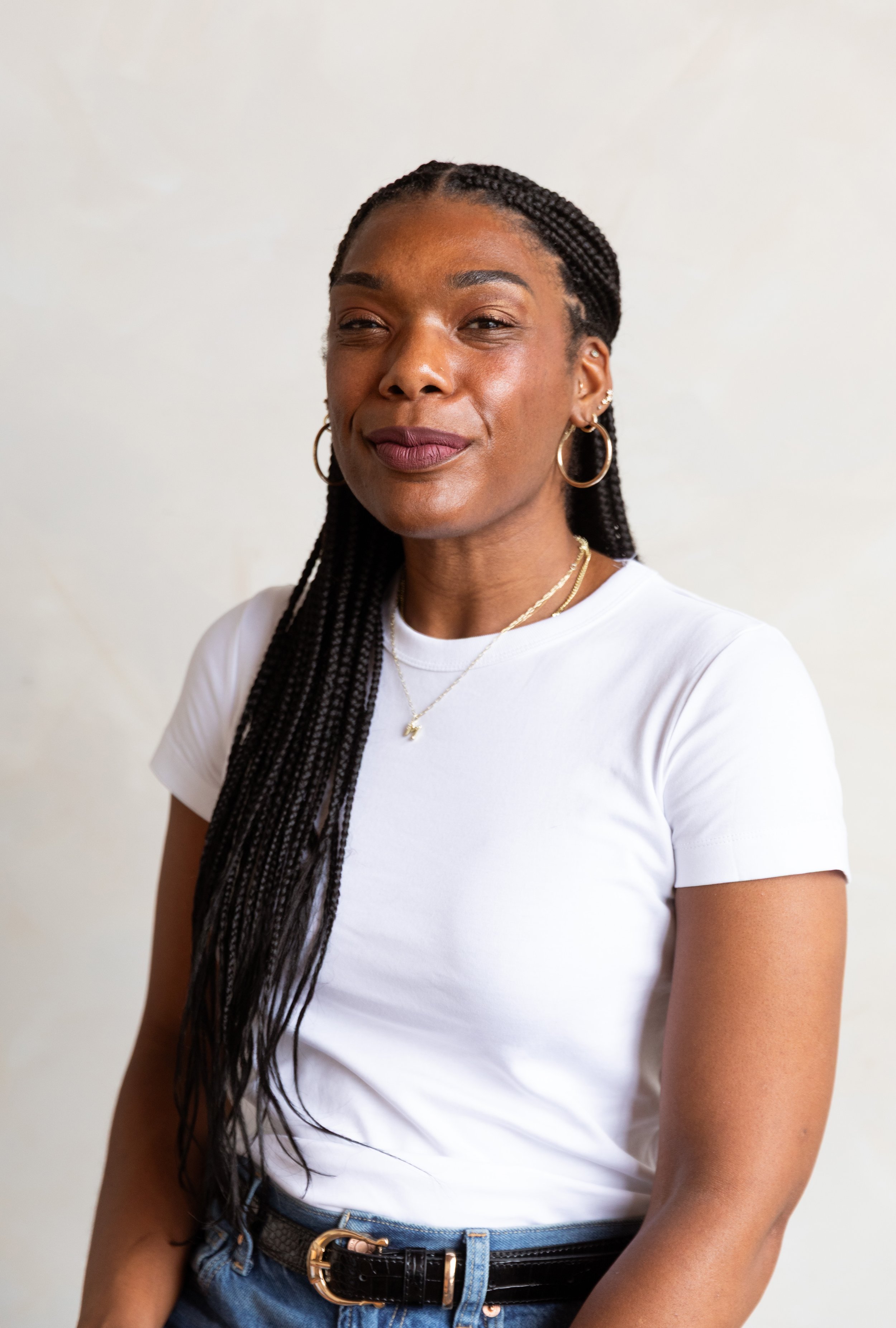 A woman with braided hair, hoop earrings, and layered necklaces wearing a white t-shirt, blue jeans, and a black belt with a gold buckle, smiling slightly against a plain light-colored background.