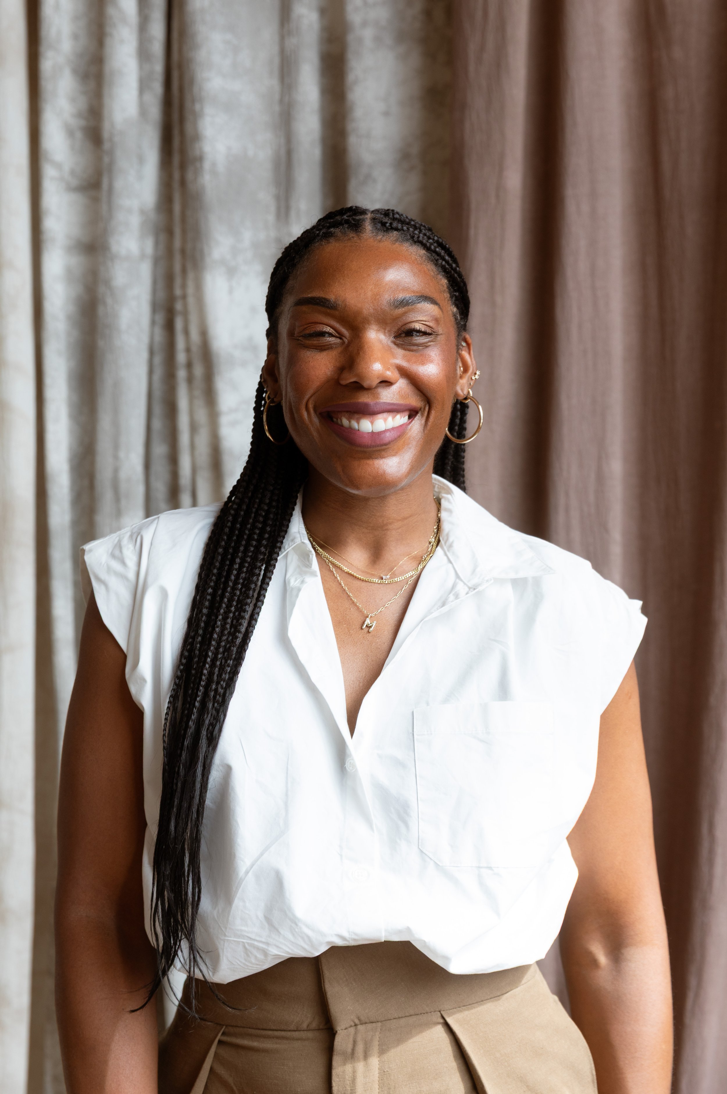Young woman smiling, wearing a white shirt, gold hoop earrings, layered necklaces, and beige pants, standing in front of brown curtains.