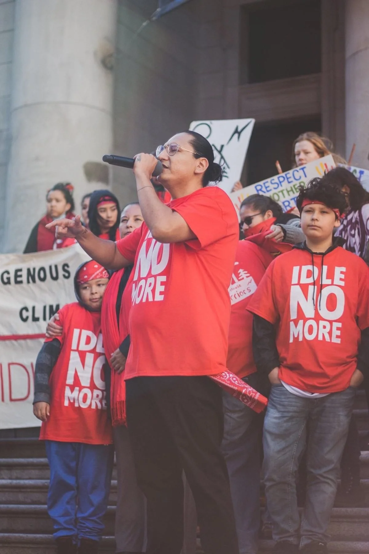 Dakota Bear speaking on the mic in an Idle No More shirt on the Vancouver Art Gallery steps, surrounded by youth