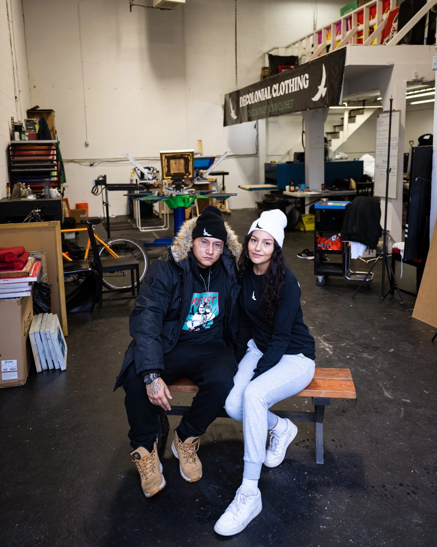 Dakota Bear and Casey Desjarlais in the Decolonial Clothing print shop in Burnaby, with the screen printing press behind them