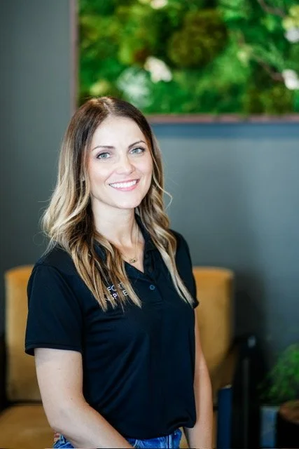 A woman with long blonde hair wearing a black polo shirt, smiling indoors with a green and white abstract art piece on the wall behind her.
