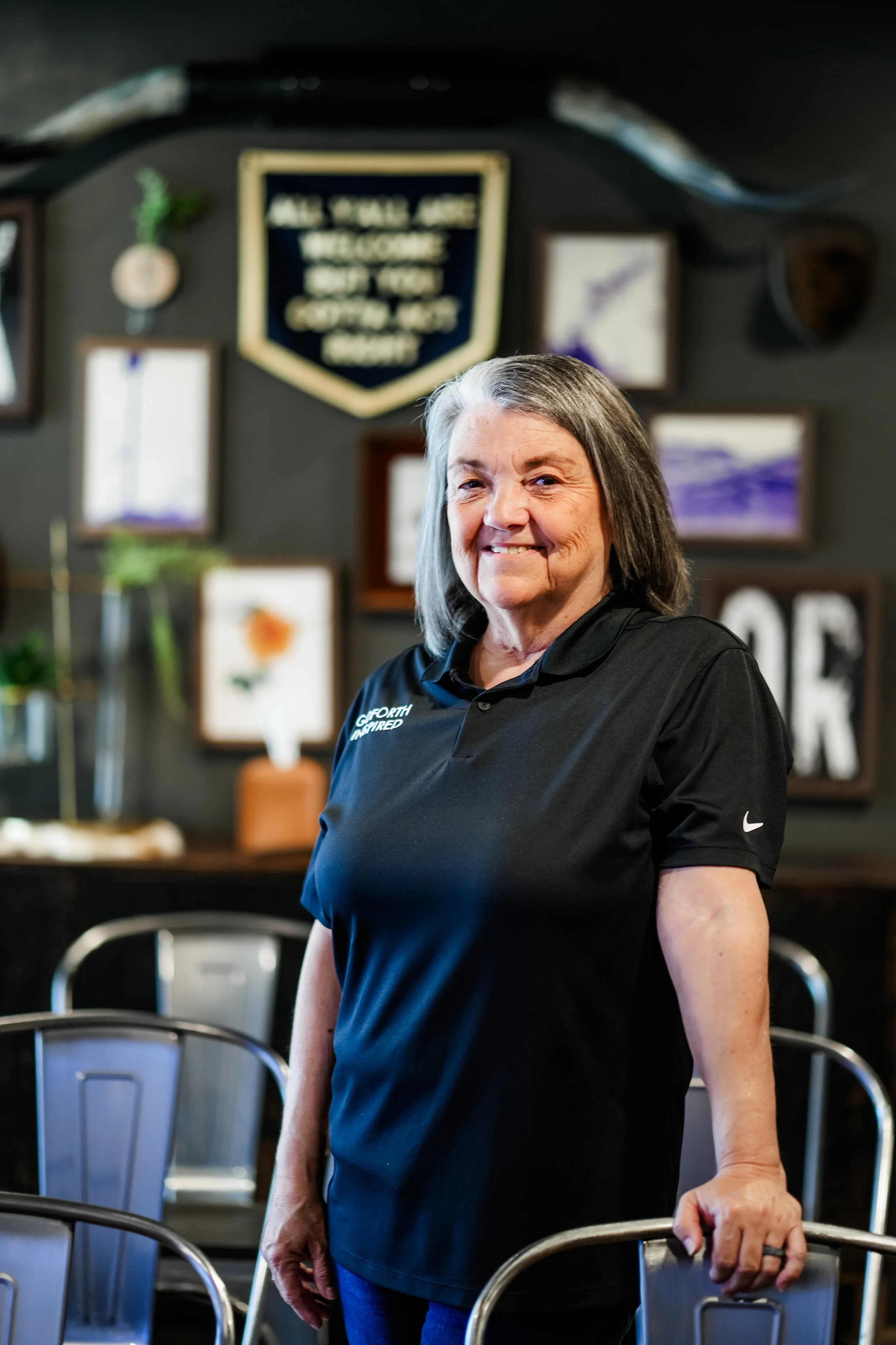 An older woman with gray hair wearing a black polo shirt and smiling, standing in a dining area with chairs and framed artwork on the wall behind her.