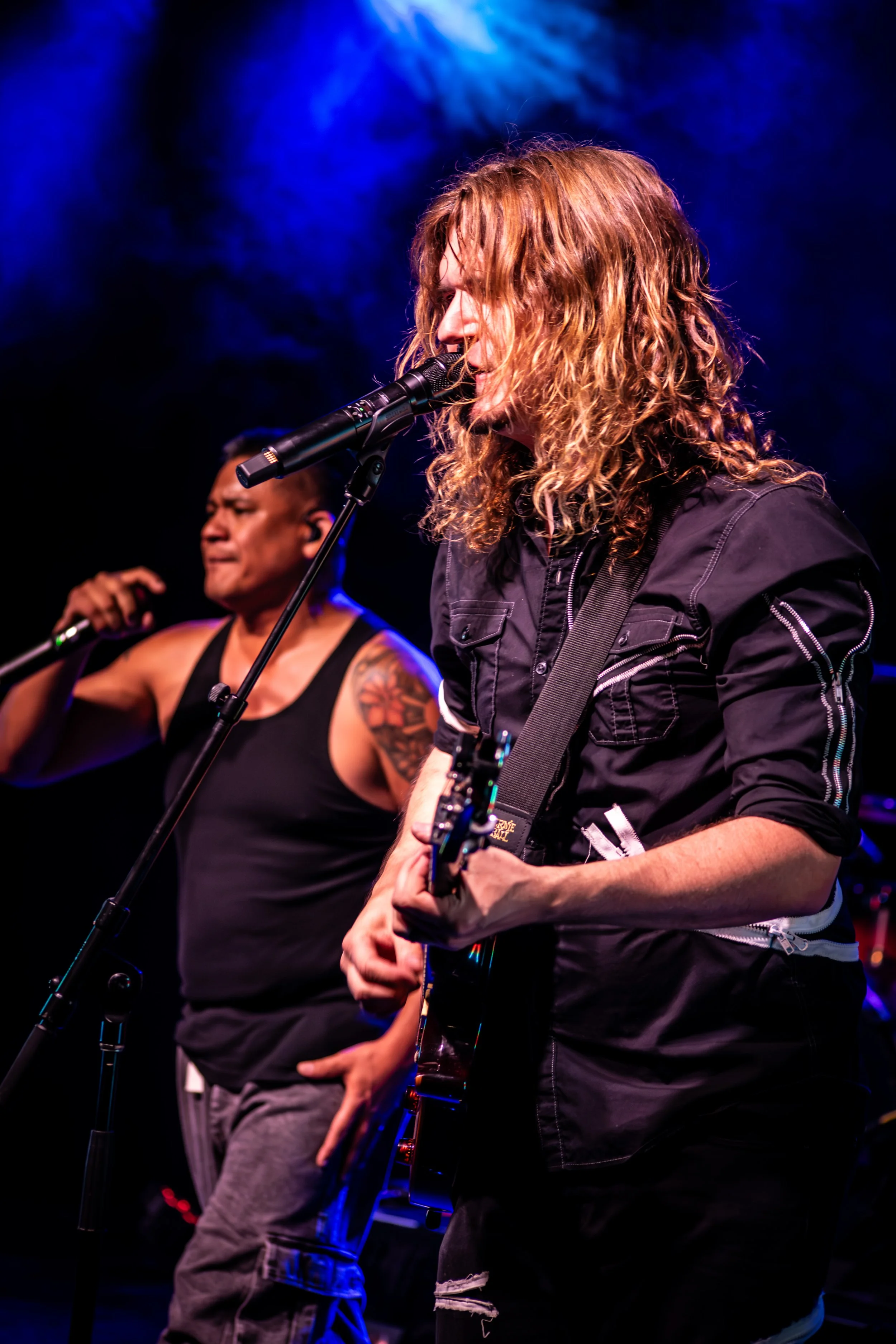 Male singer and guitarist performing on stage under blue lighting, with a microphone in front of him.
