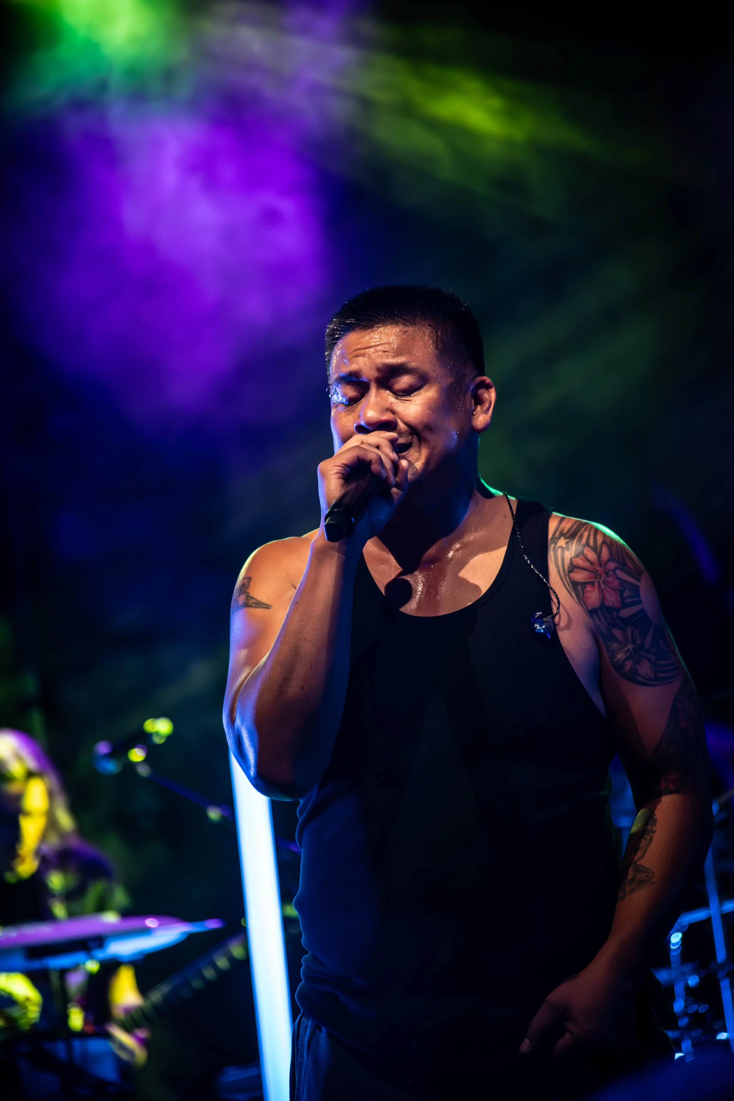 A man with tattoos on his arms and neck, wearing a black tank top, passionately singing into a microphone on stage under colorful purple, green, and blue lights.