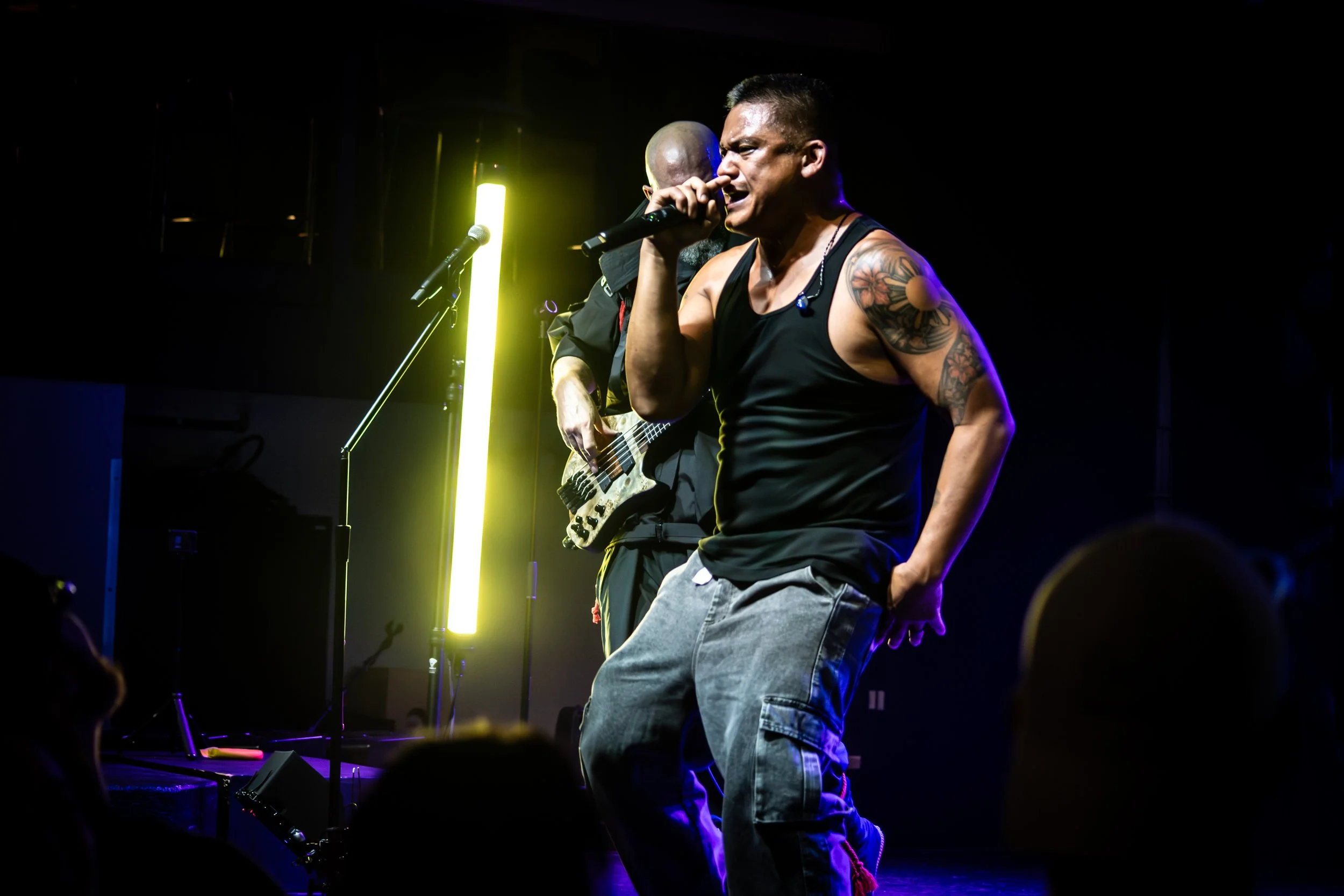 A male performer singing into a microphone on stage, wearing a black tank top and jeans, with tattoos on his arm, illuminated by stage lights.