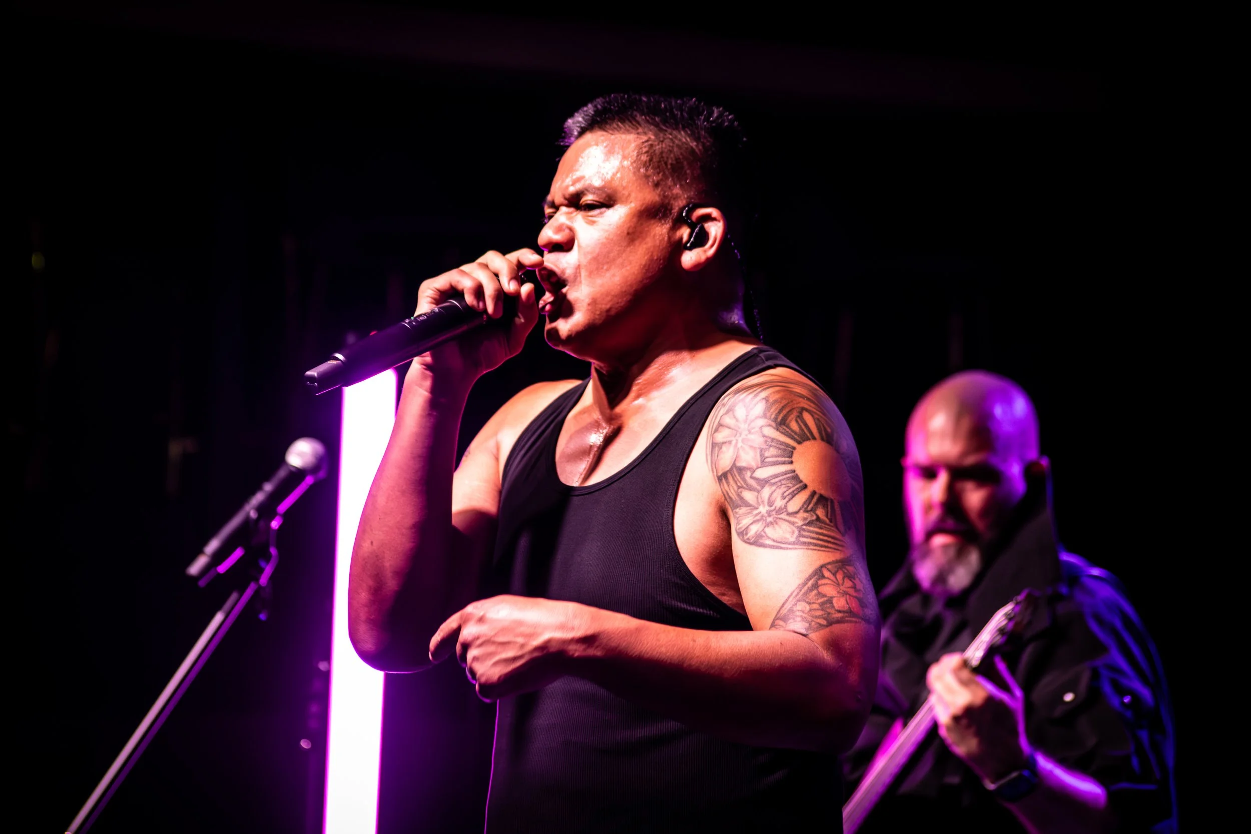 A male singer with tattoos on his arm, holding a microphone and singing on stage with purple lighting, with a band member playing guitar in the background.