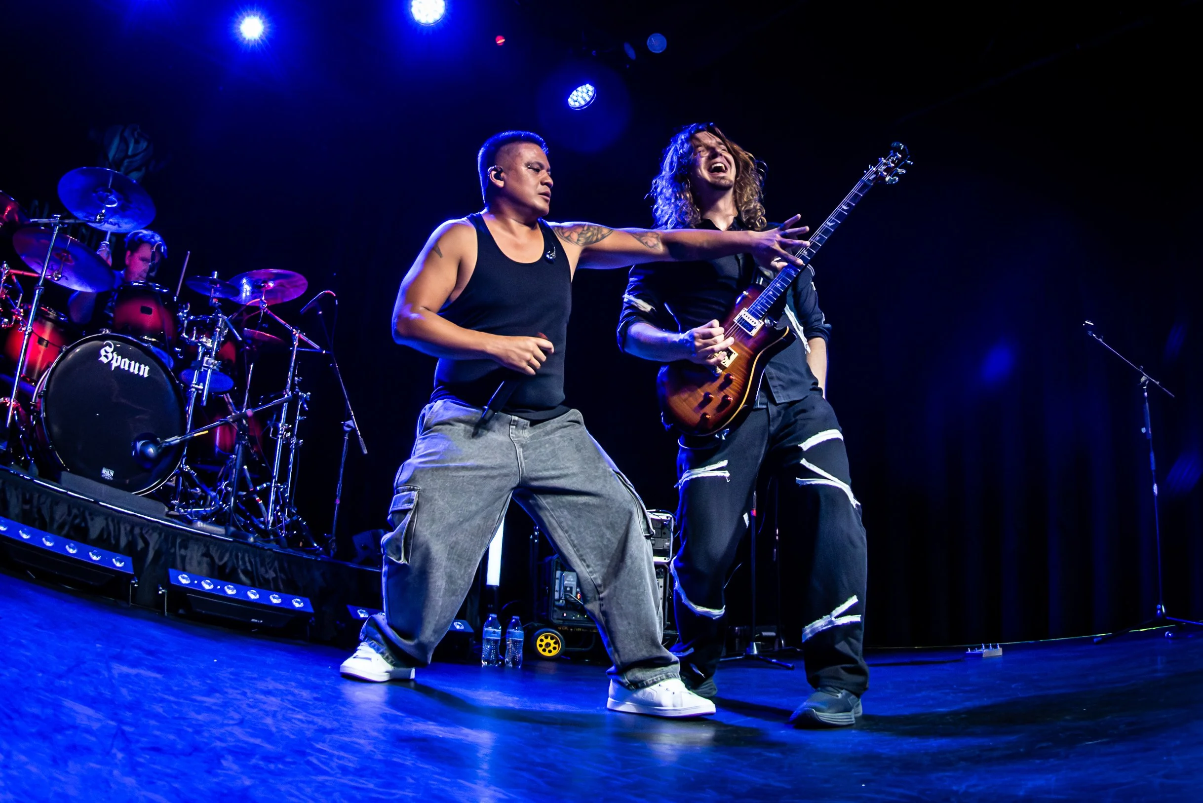 Two male musicians perform on stage with a drummer in the background. One is wearing a black sleeveless shirt and baggy pants, and the other is playing a guitar, wearing a black outfit with white stripes at the knees. The stage is lit with blue and w