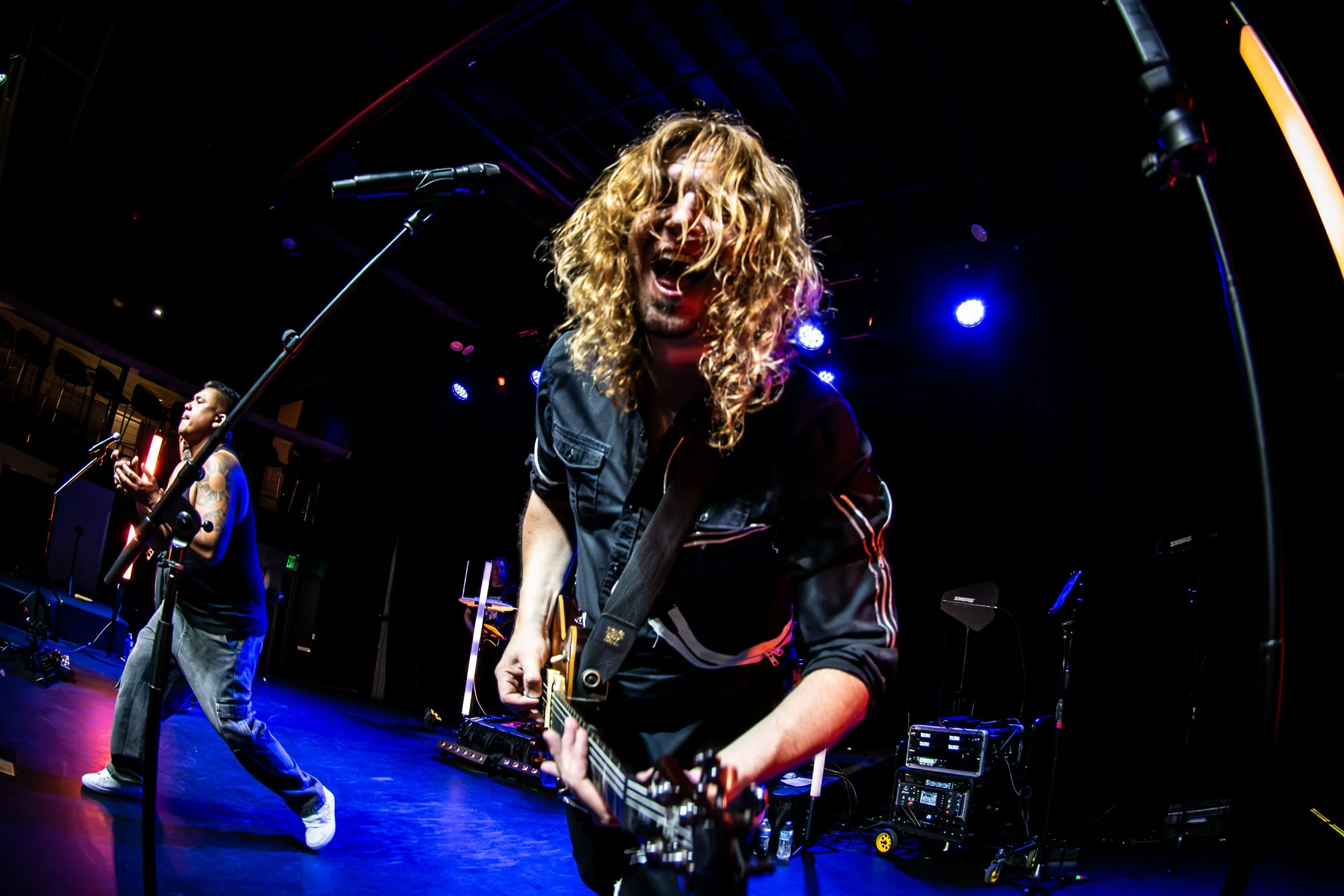 Musicians performing on stage with colorful lighting, one playing an electric guitar and the other singing into a microphone.