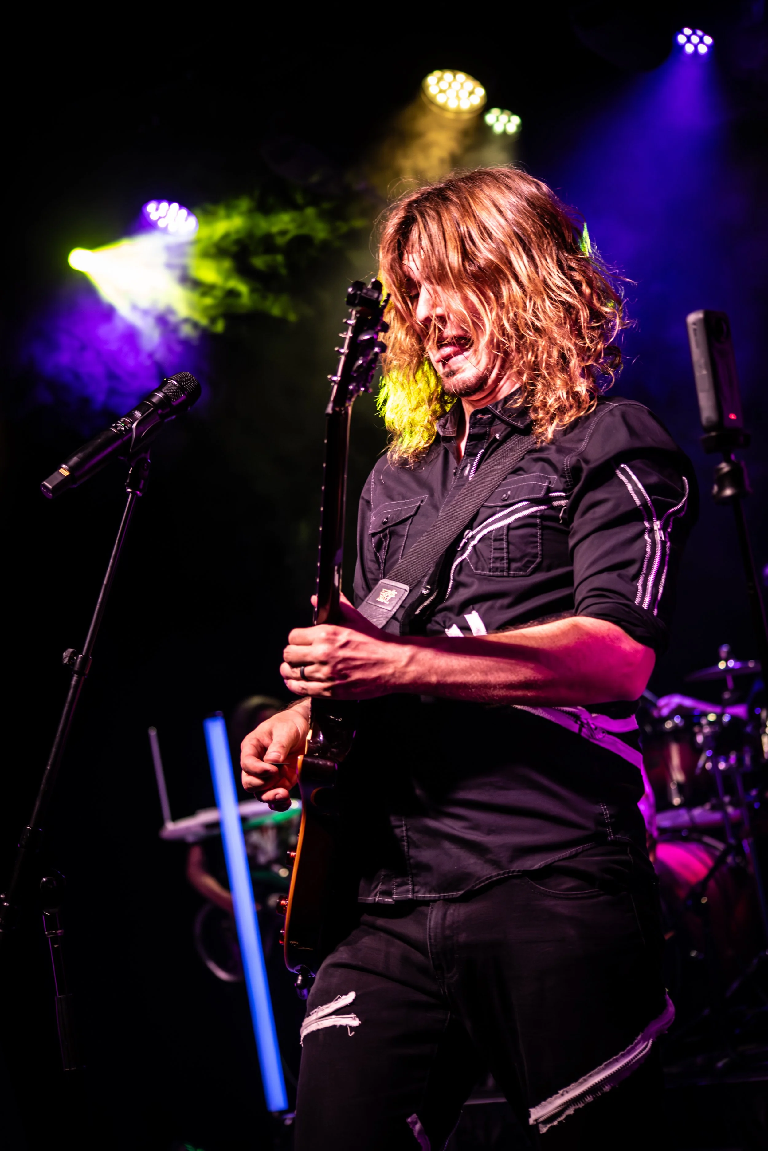 Musician with long curly hair playing an electric guitar on stage with colorful stage lighting.