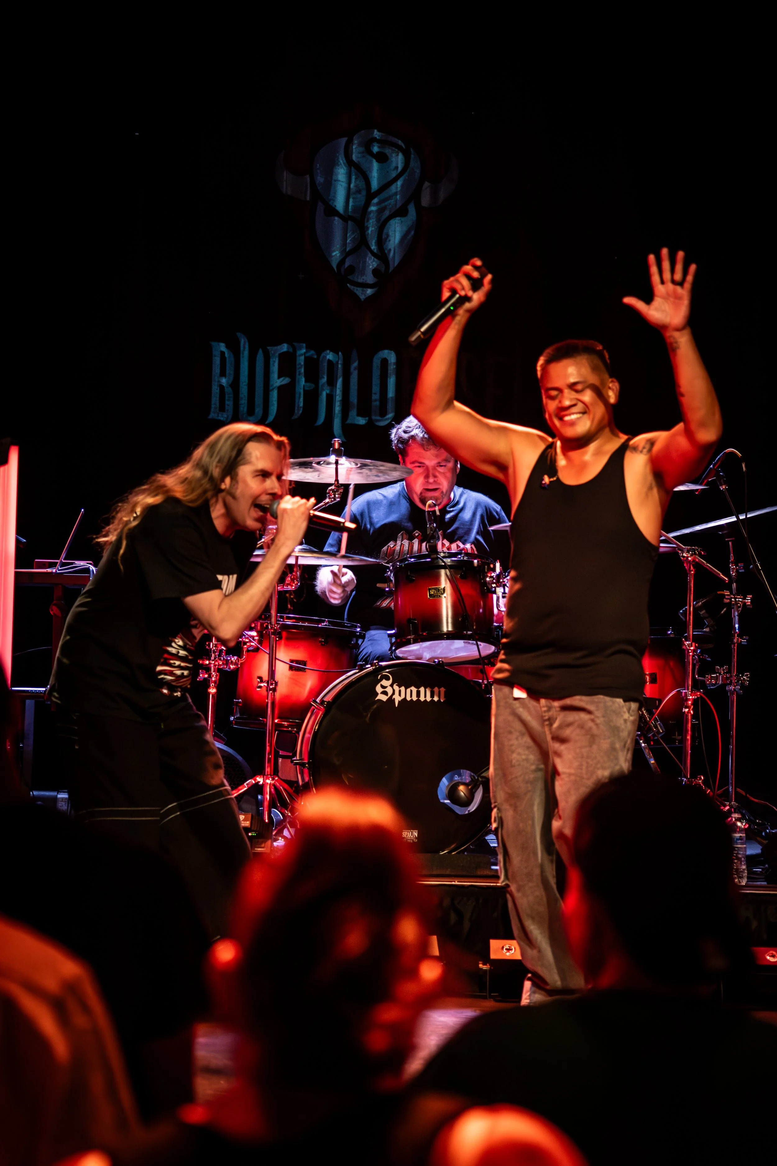 A live musical performance featuring three band members on stage. A singer with long hair and a microphone, a man playing the drums, and another man with short hair and tattoos, raising his hand and smiling. The backdrop displays the word 'BUFFALO' a