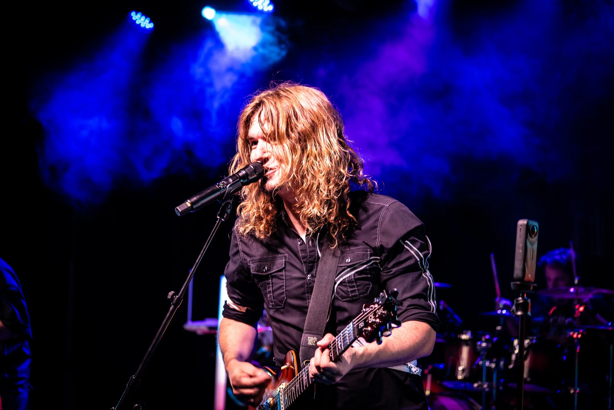 A woman with curly red hair singing into a microphone while playing an electric guitar on stage, with colorful stage lights and smoke behind her.