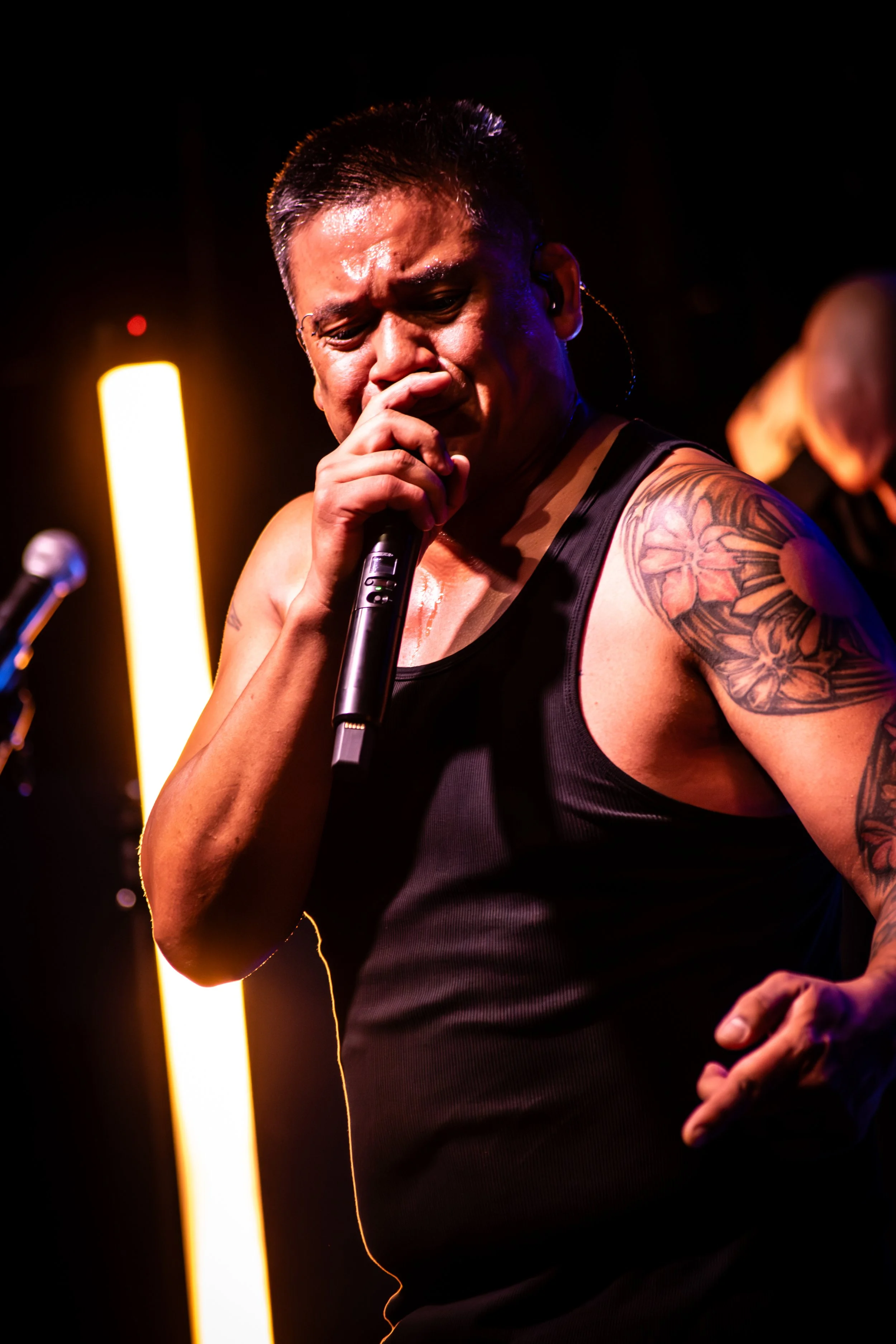 A man performing on stage, holding a microphone, sweating, with a tattooed arm, wearing a black tank top, illuminated by stage lights.