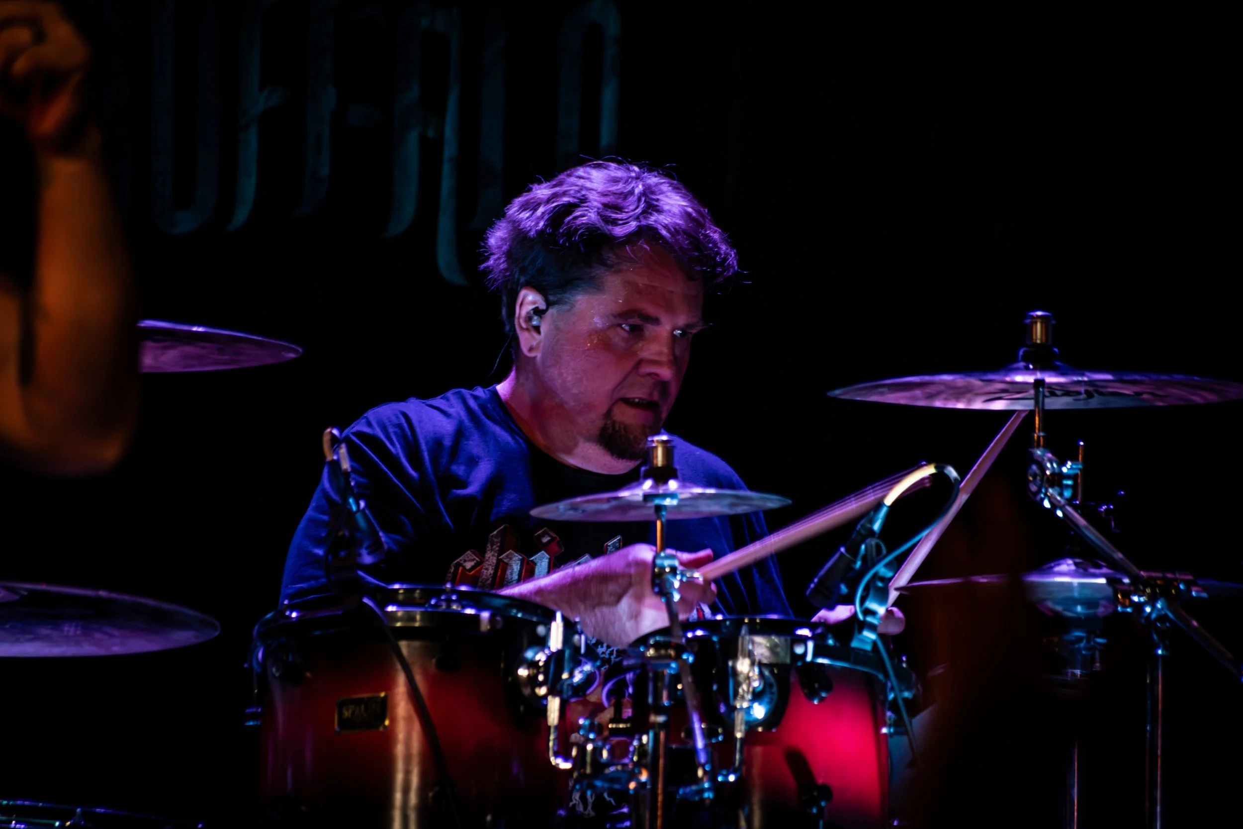 A man playing drums on stage with purple and blue lighting, focused on his performance.
