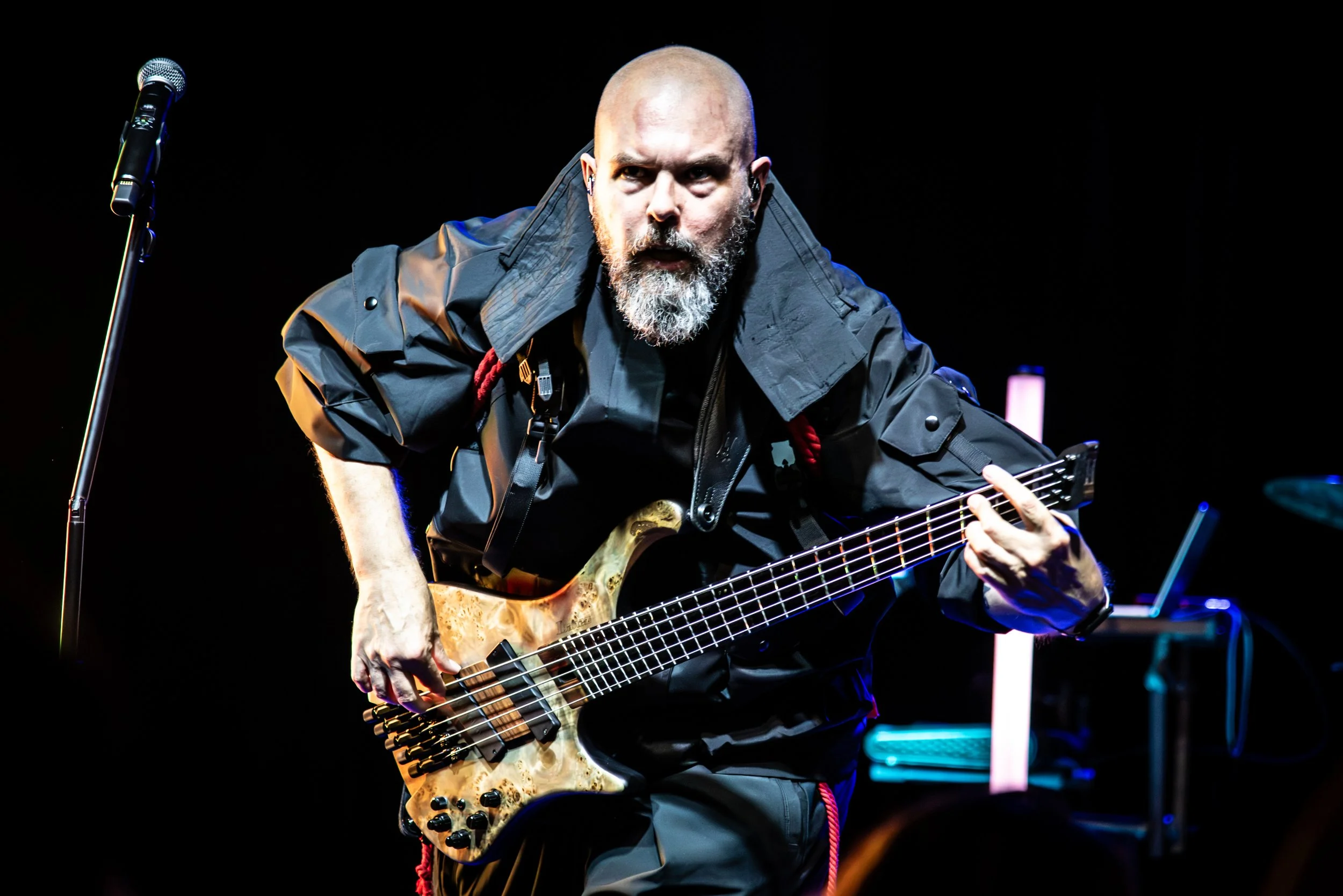 A man with a shaved head and beard playing an electric bass guitar on stage, wearing a black jacket, with a microphone stand nearby and stage lights in the background.