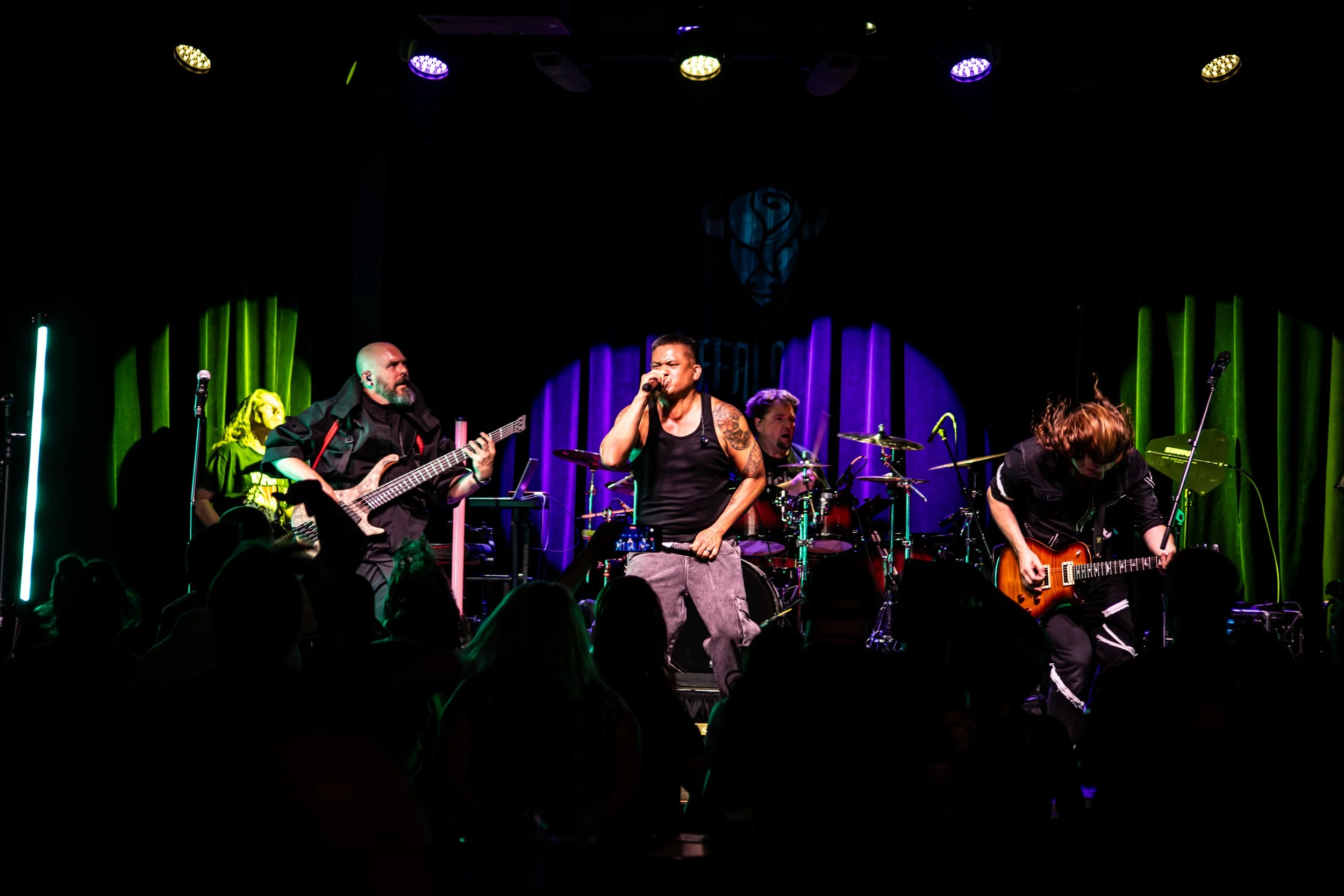 A live band performing on stage with microphones and instruments. The lead singer, a man with tattoos, is singing into a microphone. Other band members are playing guitars and drums. The stage has colorful lighting and a dark backdrop with a logo.