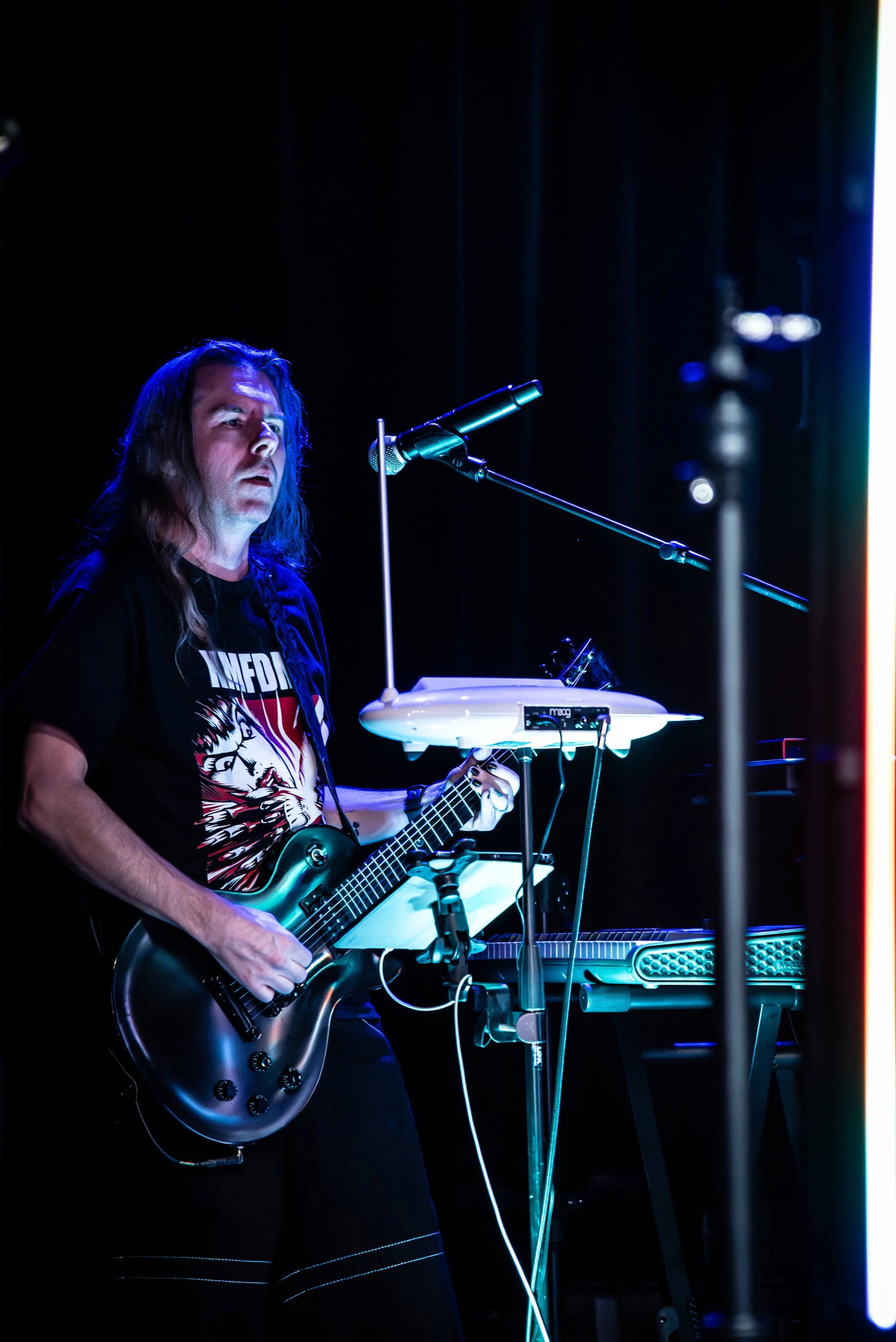 A man with long hair playing an electric guitar on stage with music equipment and a microphone, illuminated by a blue light.