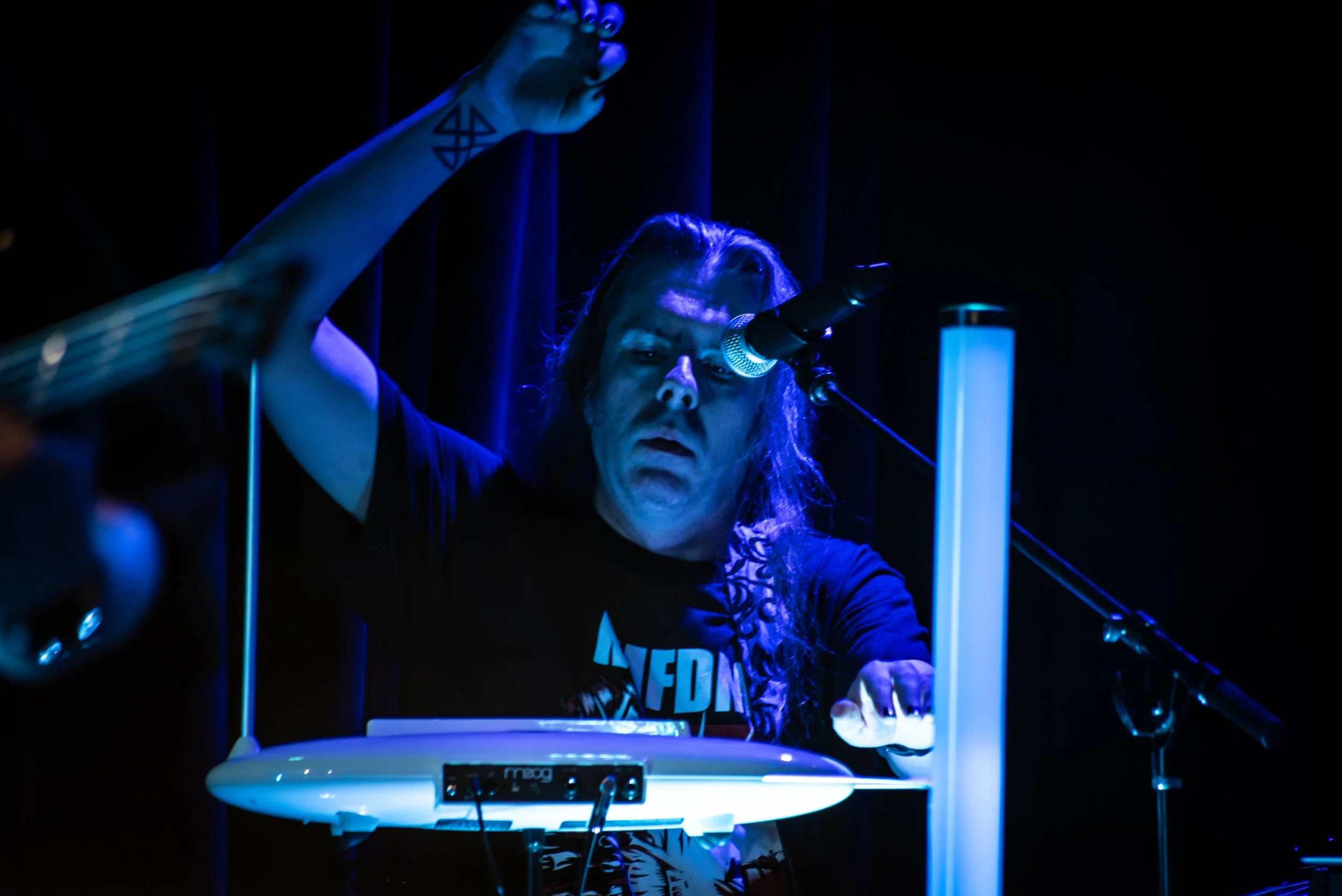 Person with long hair playing electronic musical instrument under blue lighting in a dark room.