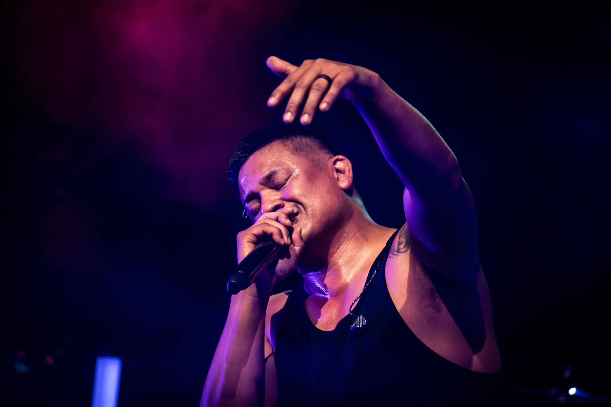 A singer with short dark hair and a tattoo on their shoulder, holding a microphone, performing on stage under purple and blue lighting, with their eyes closed and hand raised.