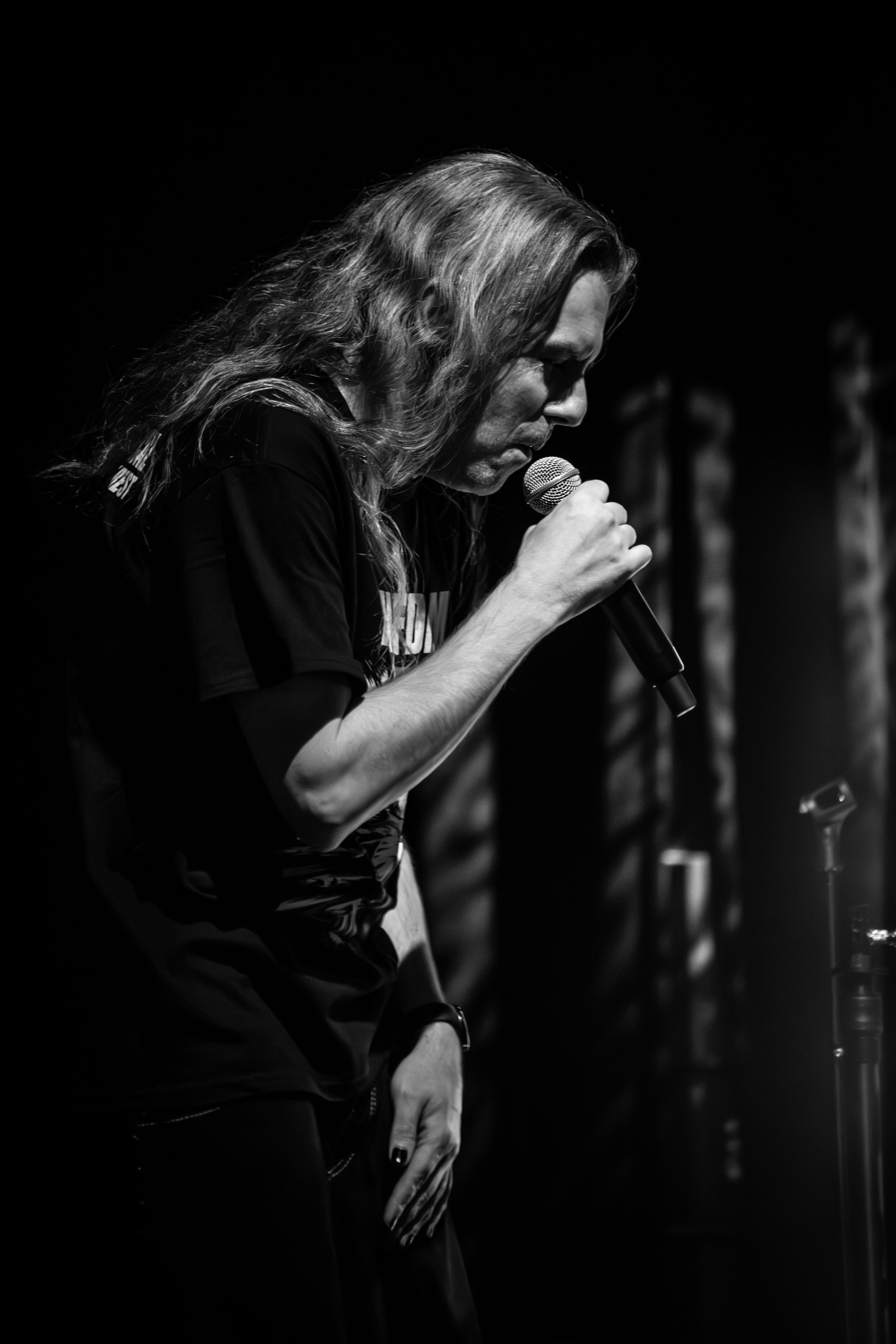 A black and white photo of a person with long, wavy hair holding a microphone, with their head tilted down and eyes closed, on a dark stage.