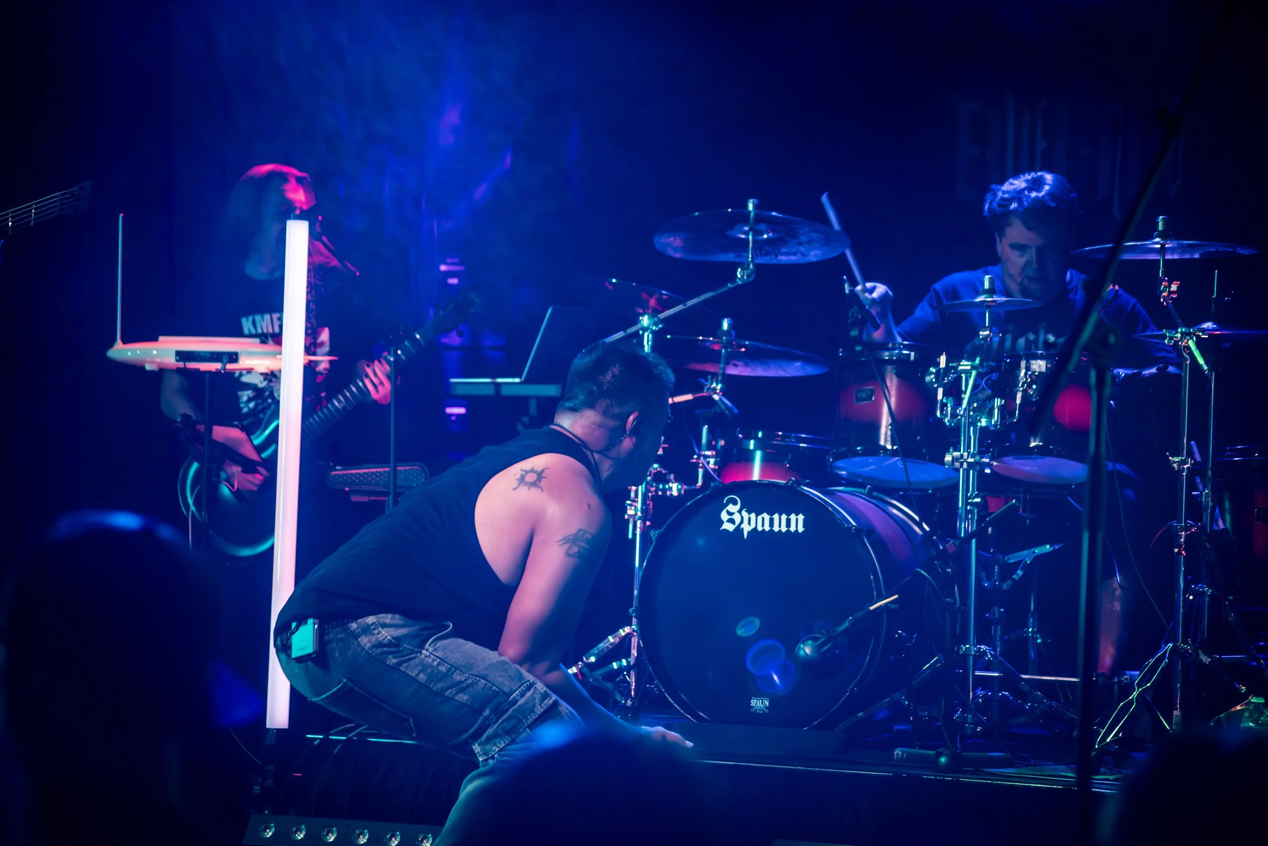 Musicians performing on stage in a dark setting with colorful lighting, featuring a guitarist, a drummer, and a keyboardist.