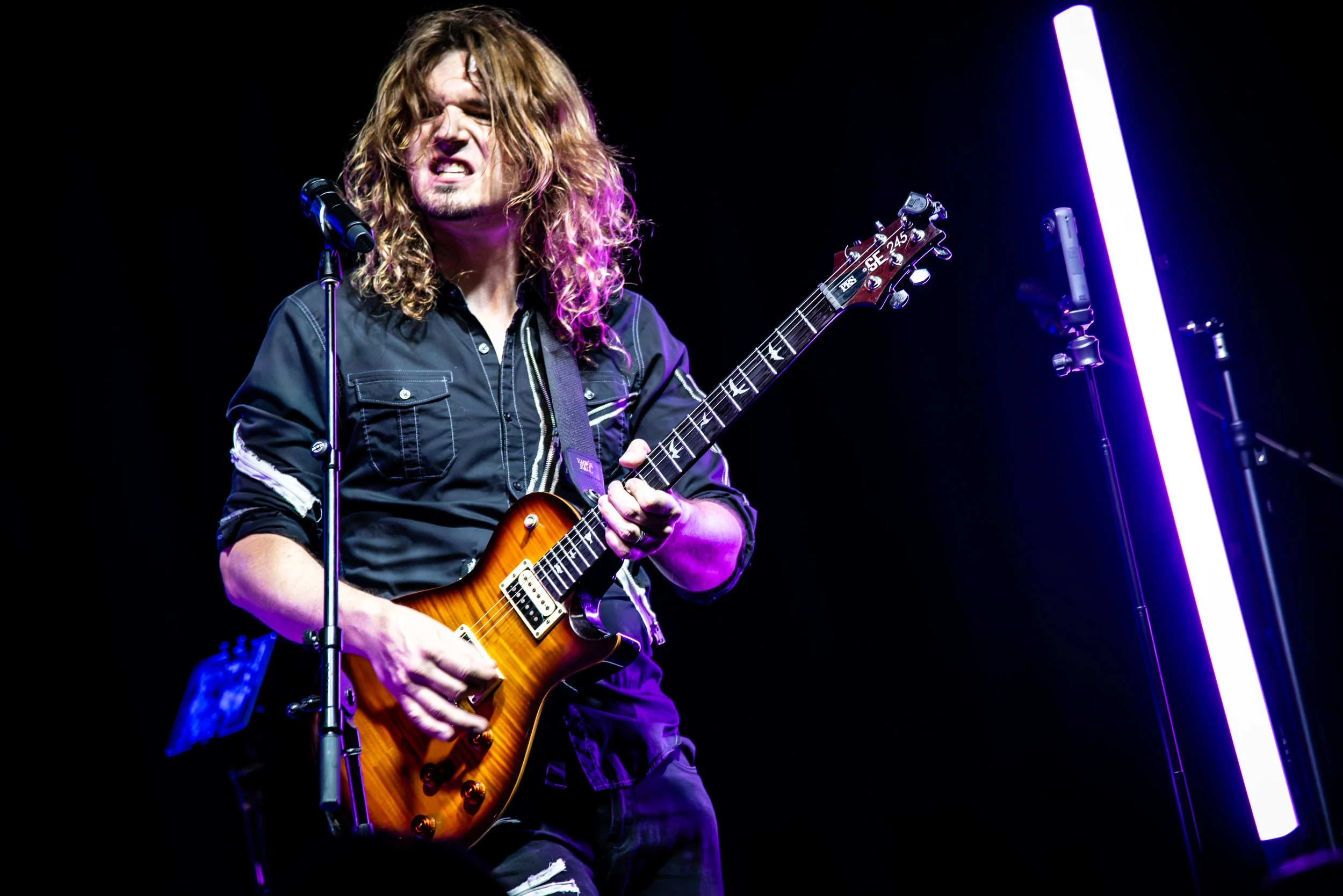 A male musician with long, wavy hair performs on stage with an orange electric guitar, wearing a dark button-up shirt, illuminated by purple and blue stage lights.