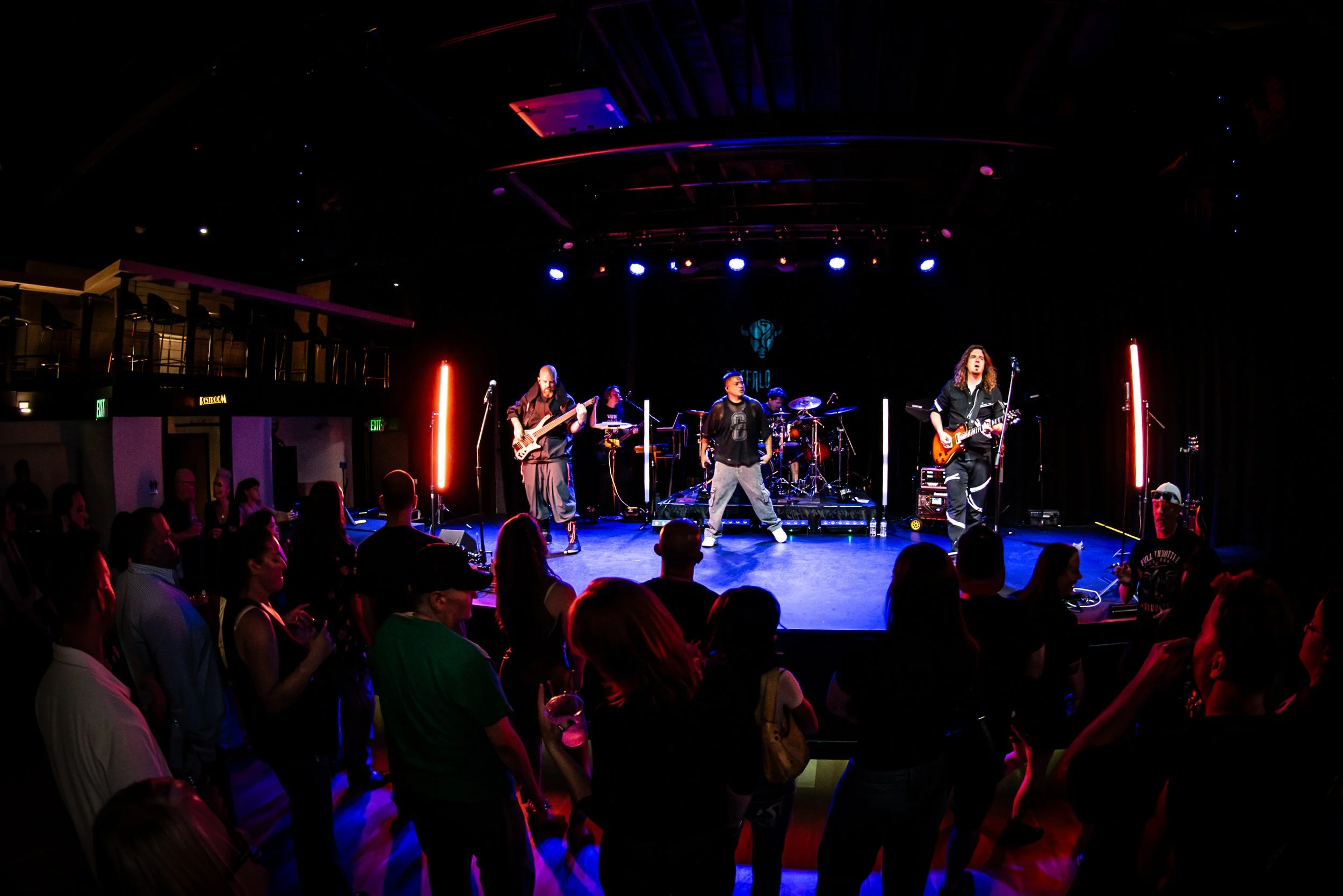 A band performing on a stage with purple and blue lighting, surrounded by an audience in a dark venue.