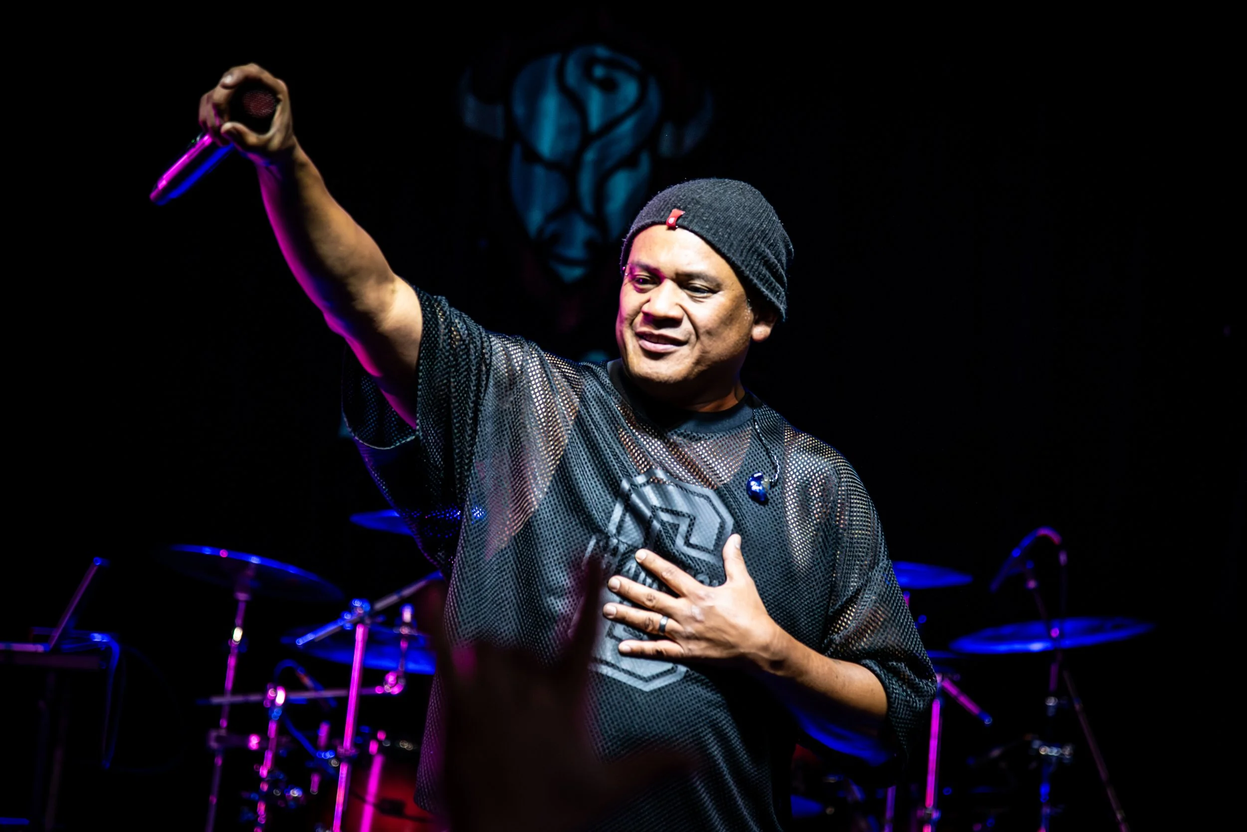 A man wearing a black beanie and a black mesh shirt is holding a microphone in one hand and pressing his other hand to his chest on stage with a dark background. There are drums in the background.