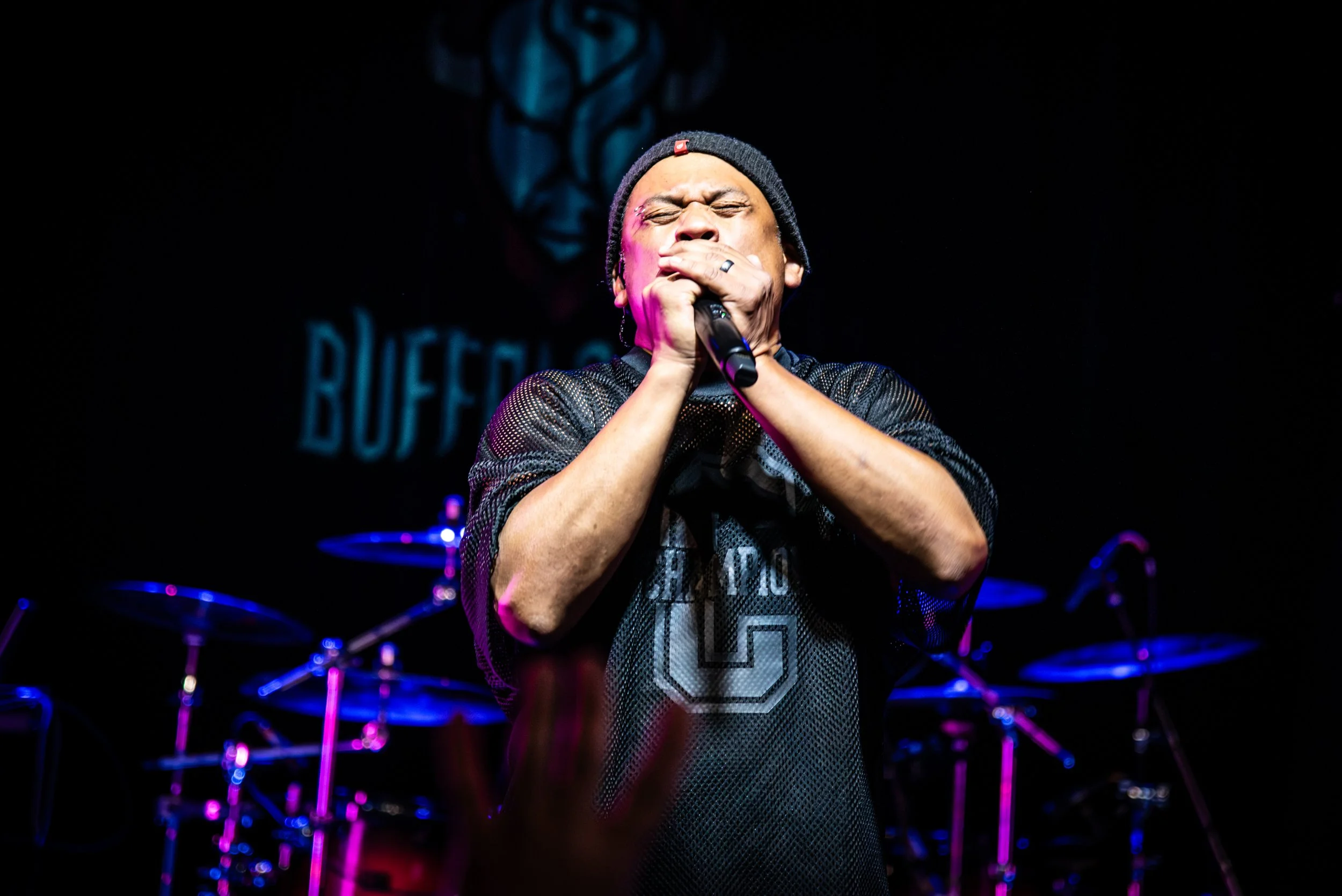 Man wearing a beanie and mesh shirt singing into a microphone on stage, with drum set and dark background.