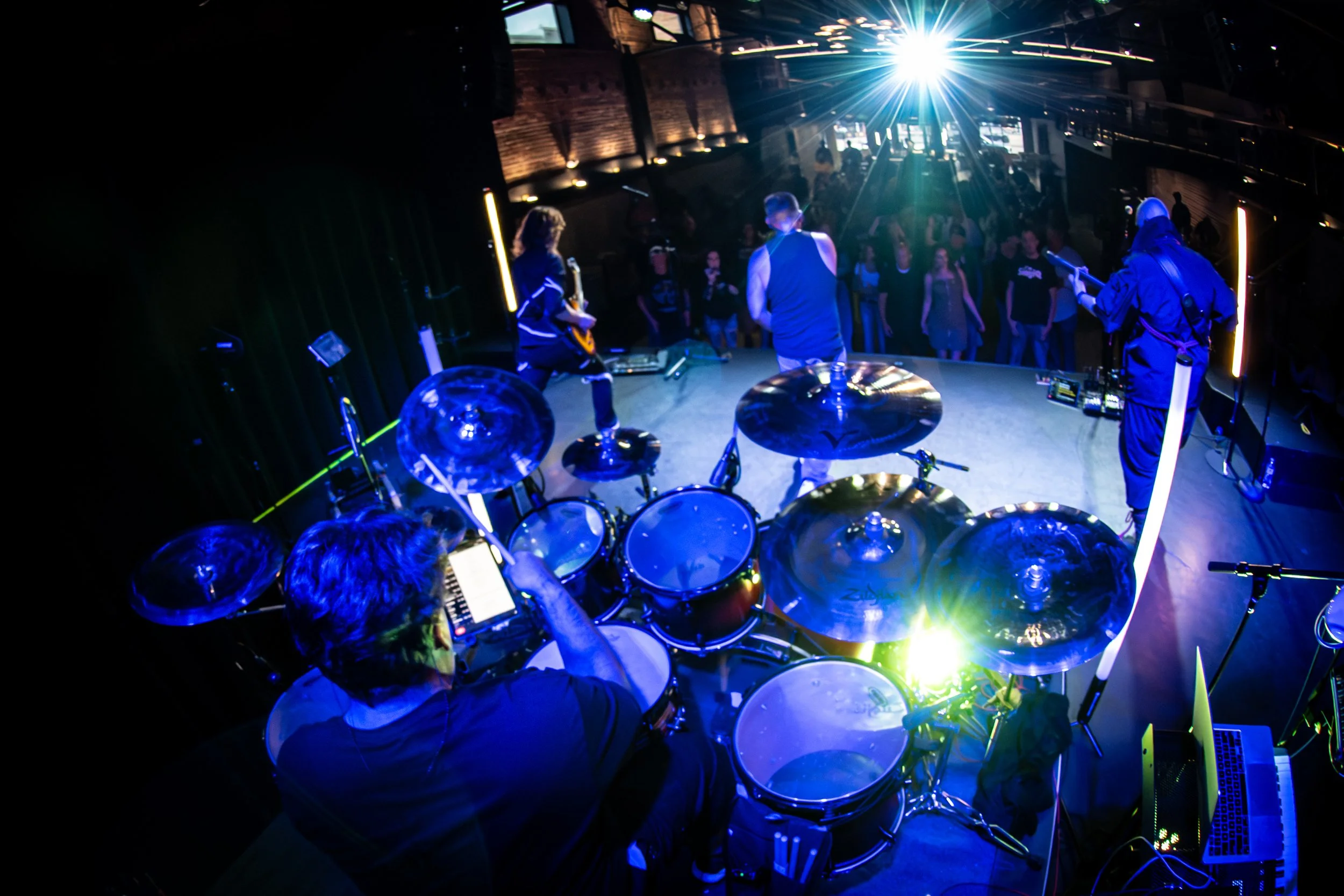 View from behind a drummer on stage with a band, audience watching, bright stage lights, and vibrant atmosphere at a music venue.