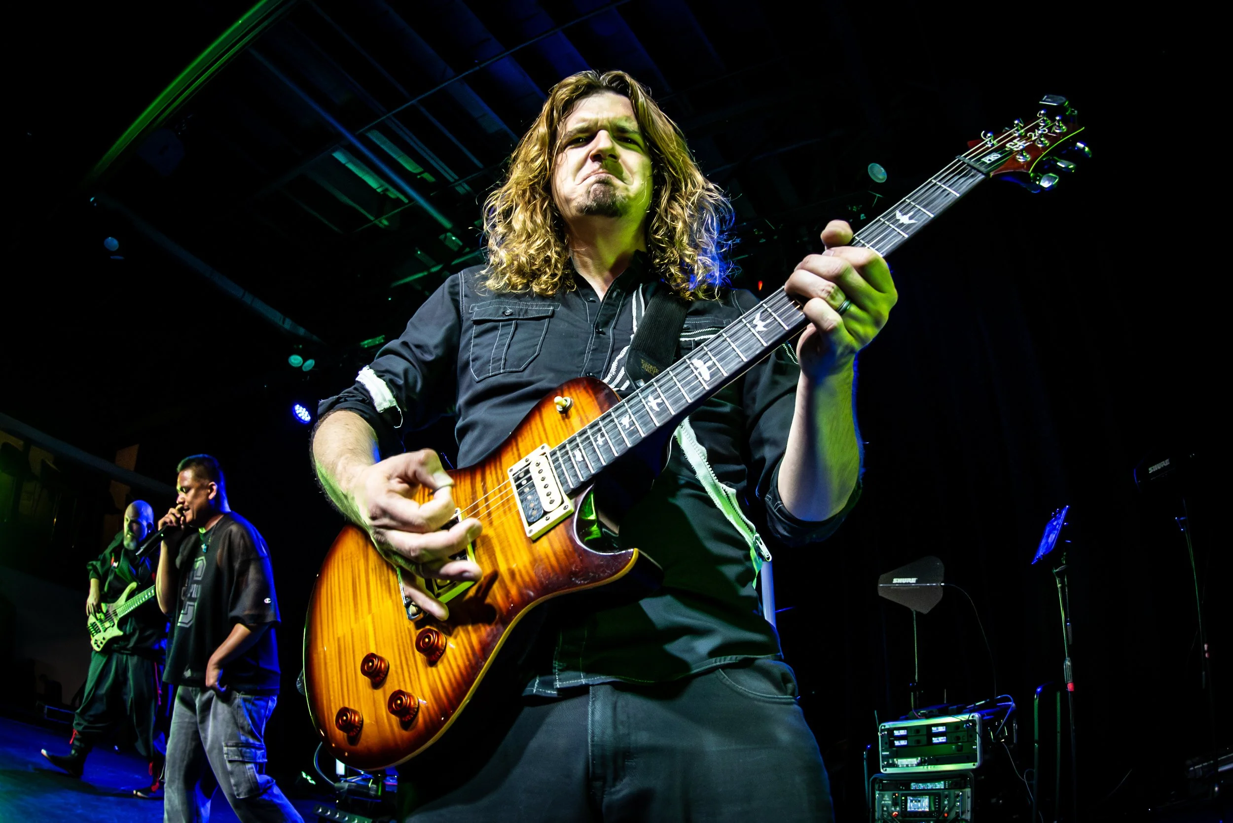 A man with long curly hair playing an orange electric guitar on stage, with band members and stage equipment visible in the background during a live musical performance.