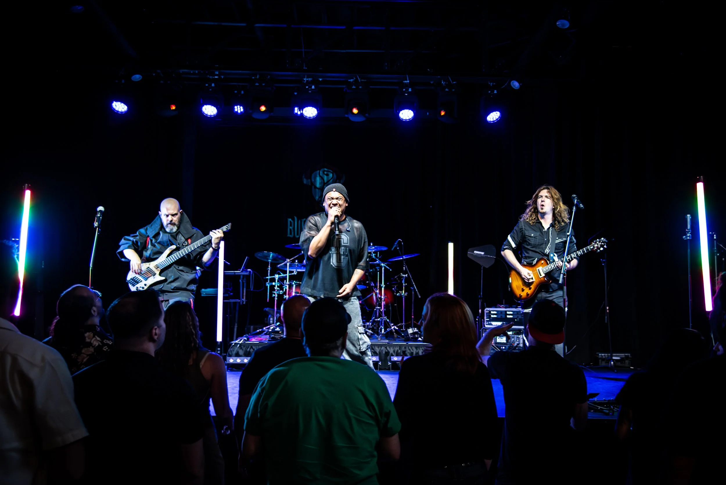 A band performing on stage with three musicians: a guitarist on the left, a singer in the center, and a guitarist on the right, with neon light columns on either side and audience members in the foreground.