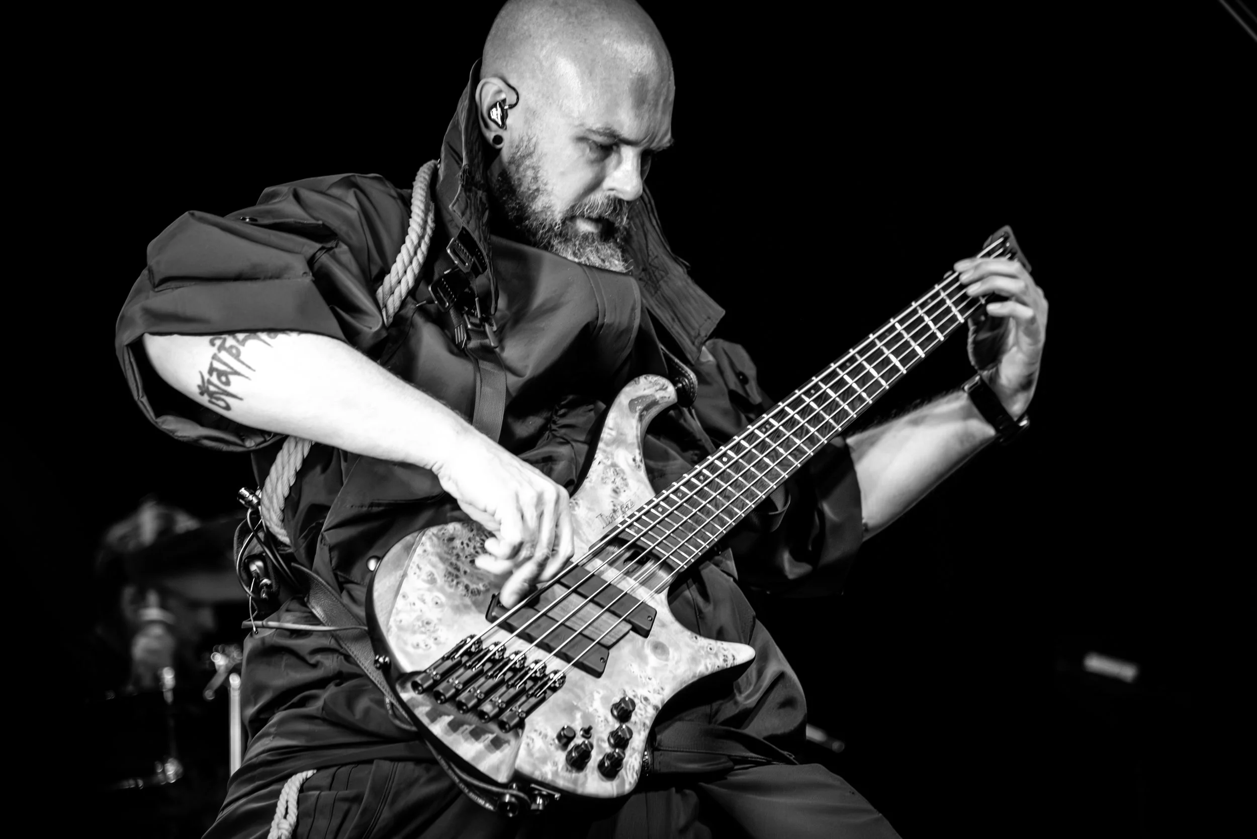 Black and white photo of a bald man with a beard playing an electric bass guitar on stage, wearing a dark shirt, with an earpiece in his ear and tattoos on his arm.