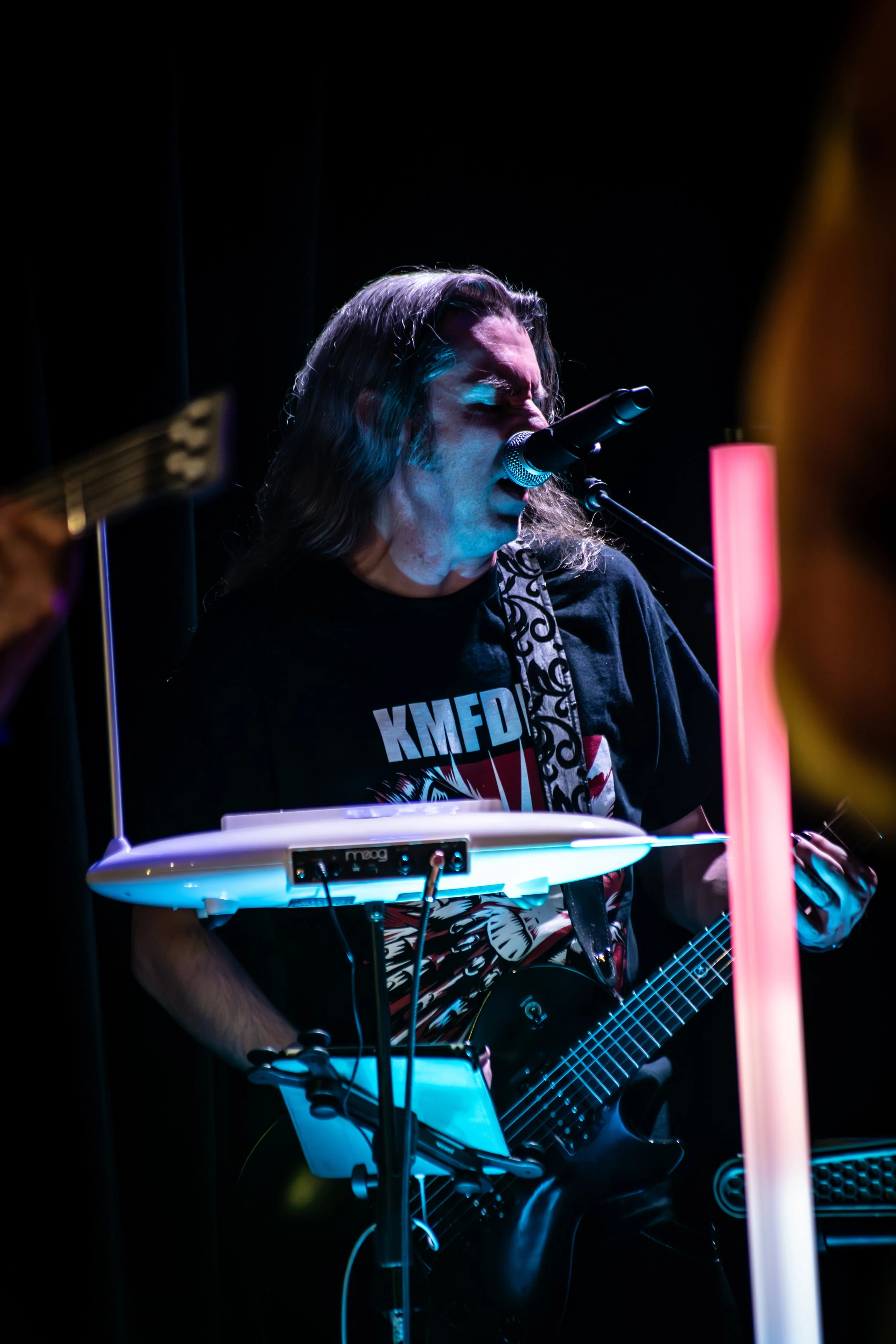 Musician performing indoors, playing guitar and singing into a microphone, with electronic equipment and colorful lighting.