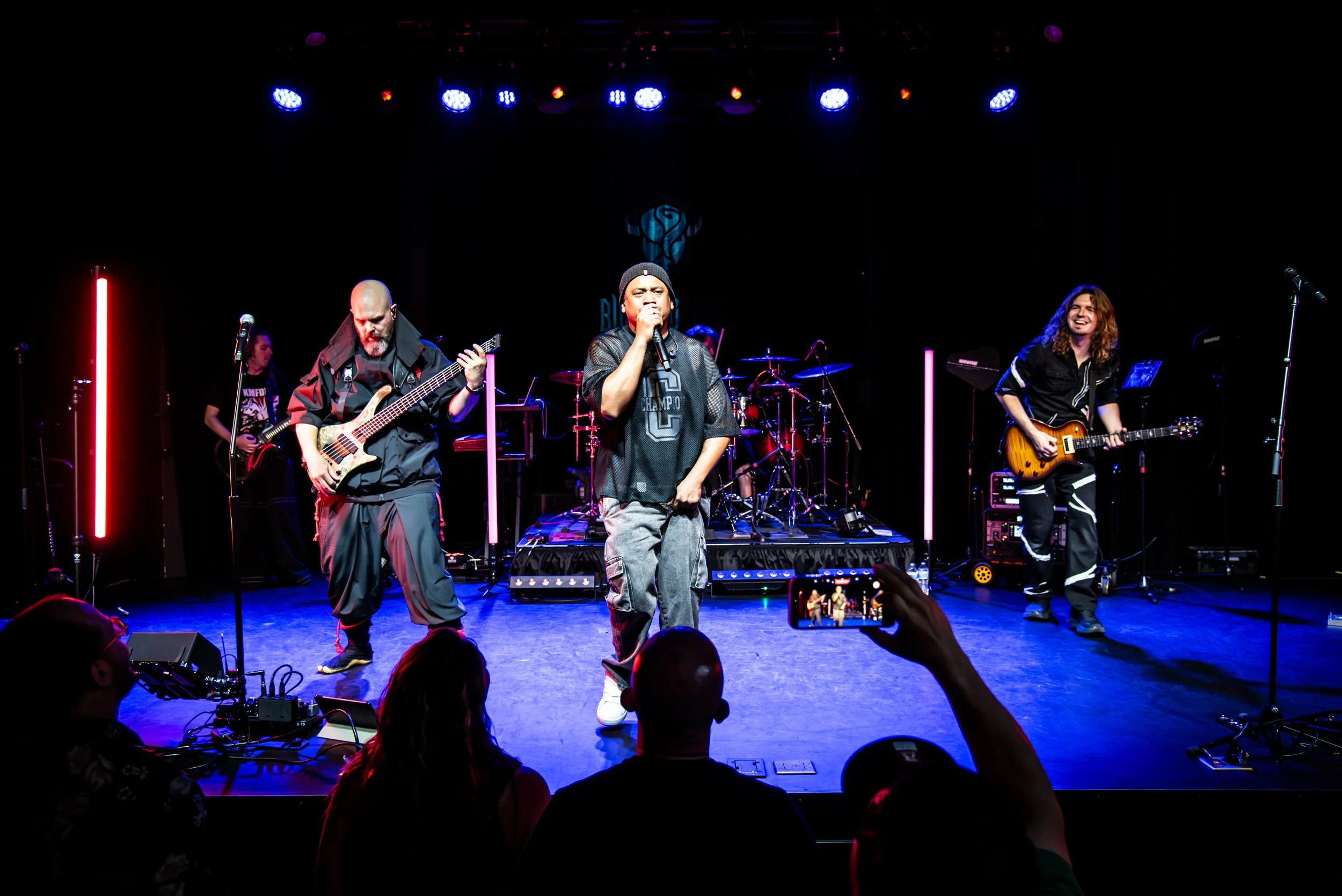Musicians performing on stage with colorful lighting, including a singer, guitarist, bassist, and drummer, with audience members taking photos.