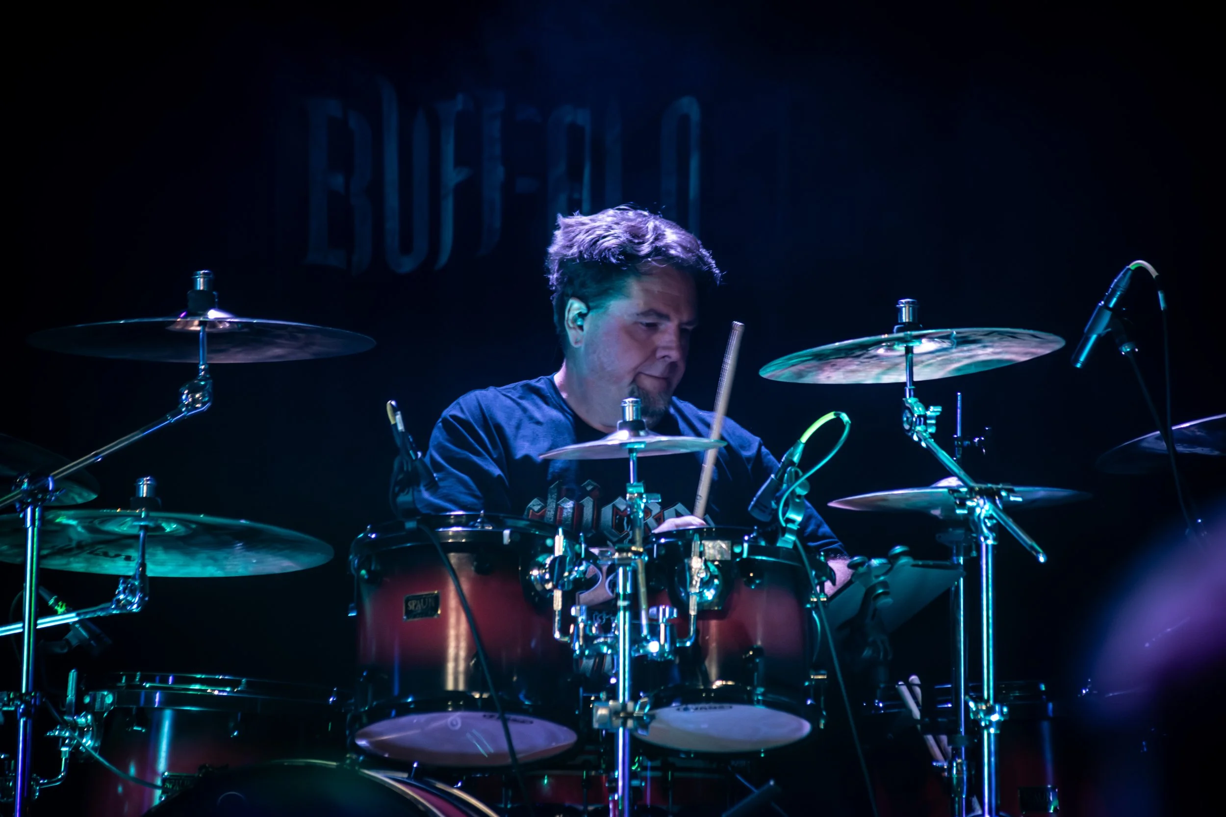 Male drummer playing a drum set on stage, with a dark background and neon lighting.