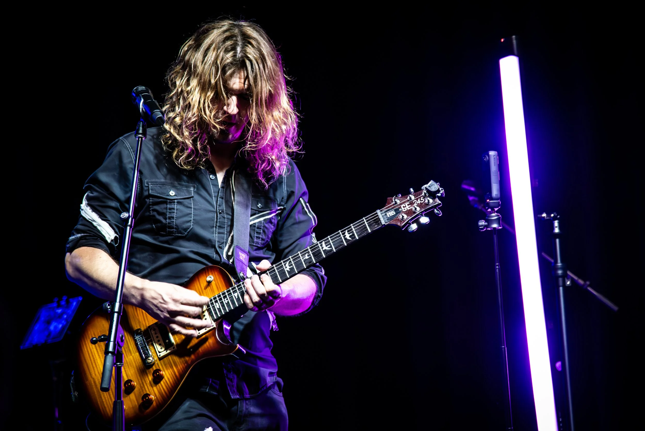 Musician playing an electric guitar on stage, illuminated by purple and white stage lights, with a microphone and stand nearby, wearing a black jacket.