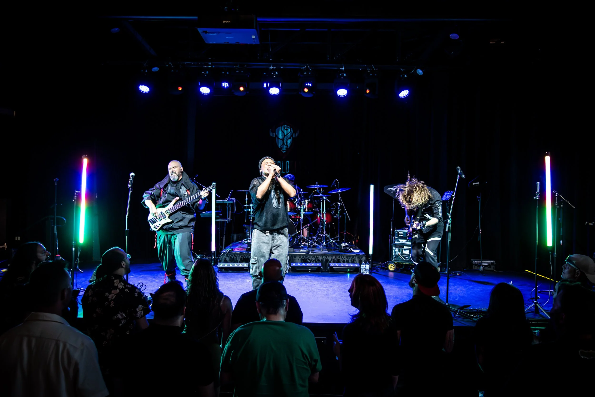 A band performs on stage with colorful vertical light pillars, with audience members watching.