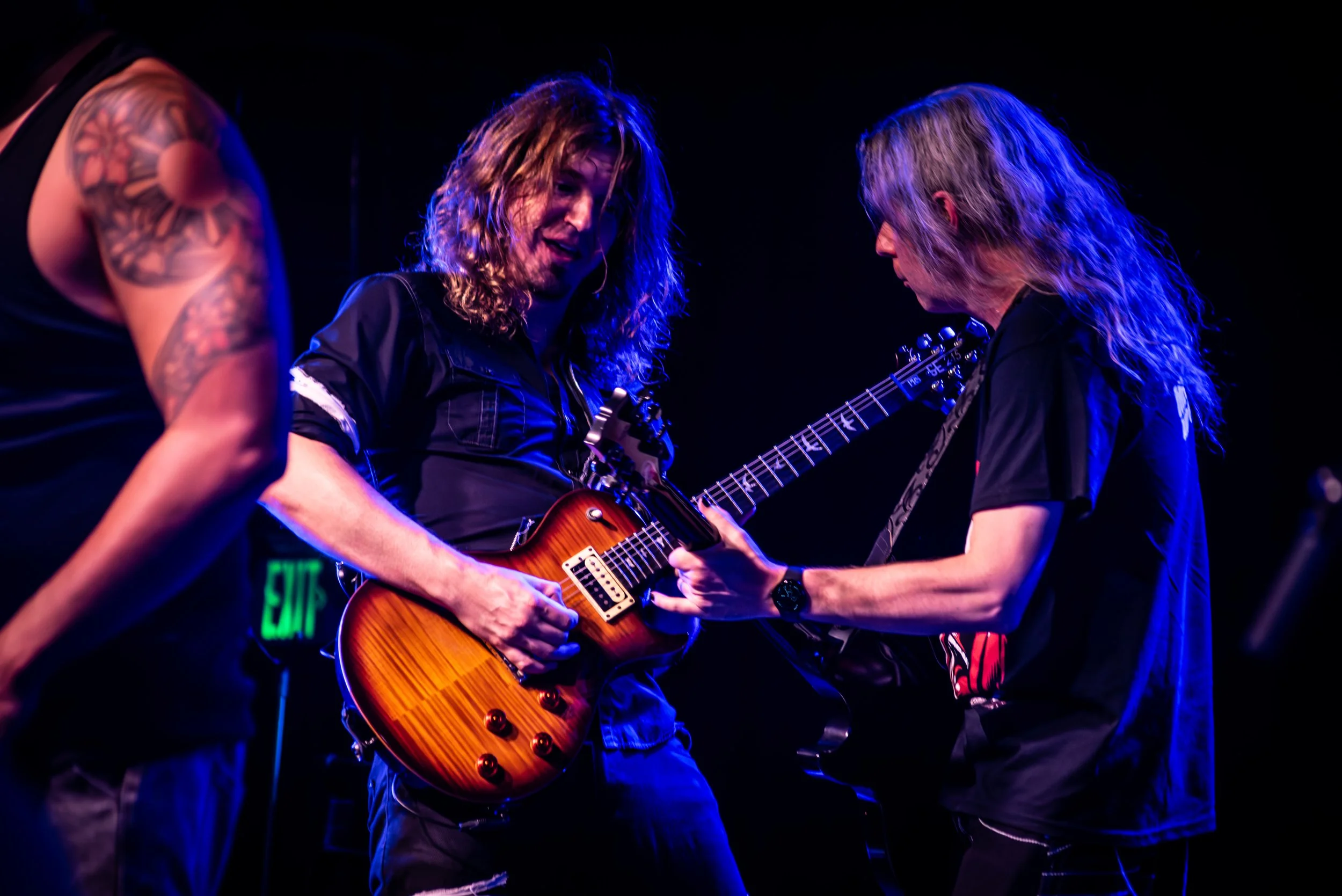 Three musicians performing on stage, one playing an electric guitar with a natural wood finish, and two others watching closely, illuminated by colorful stage lighting.