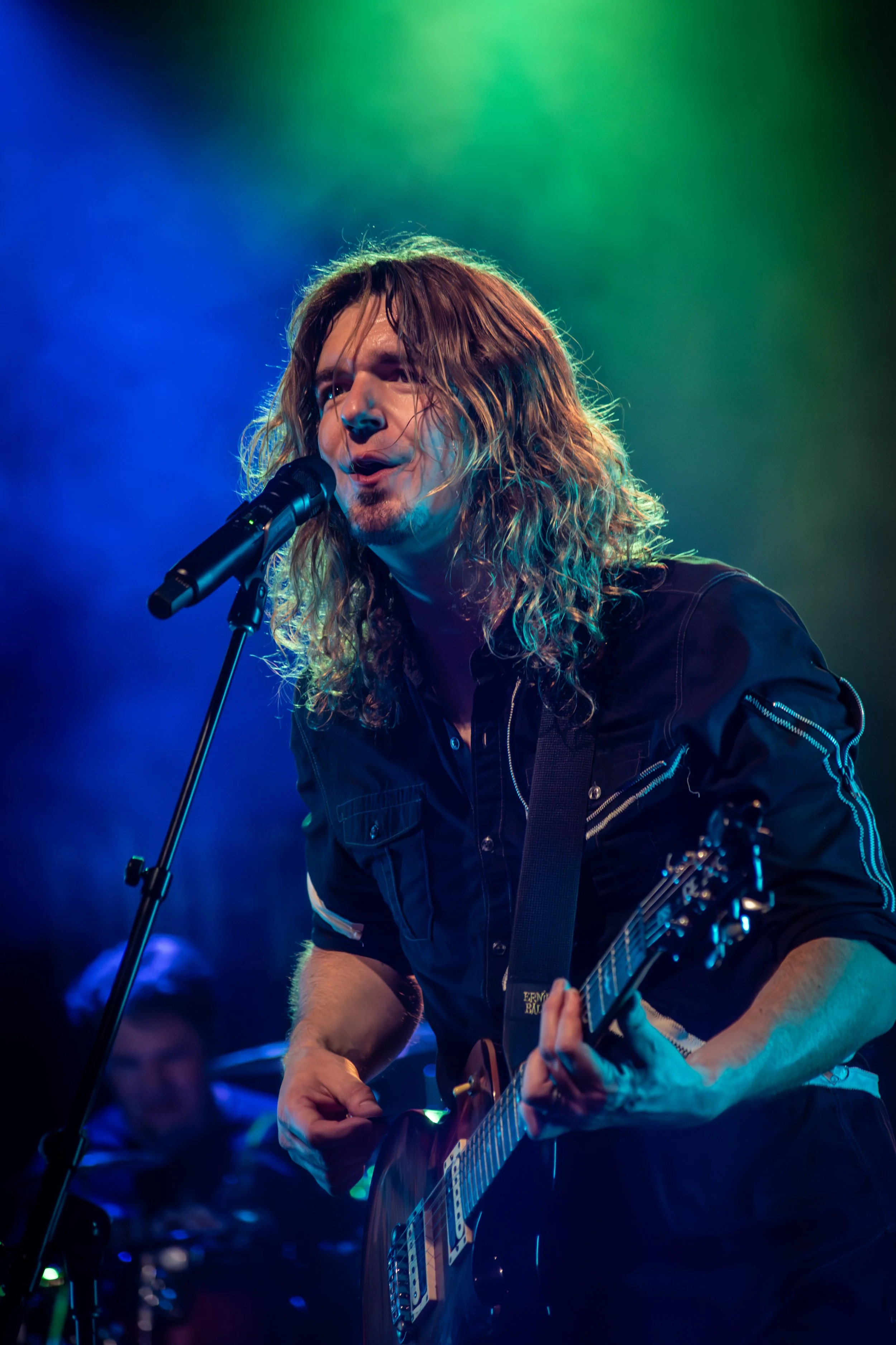Male musician with long hair playing an electric guitar on stage, singing into a microphone, under colorful stage lighting.