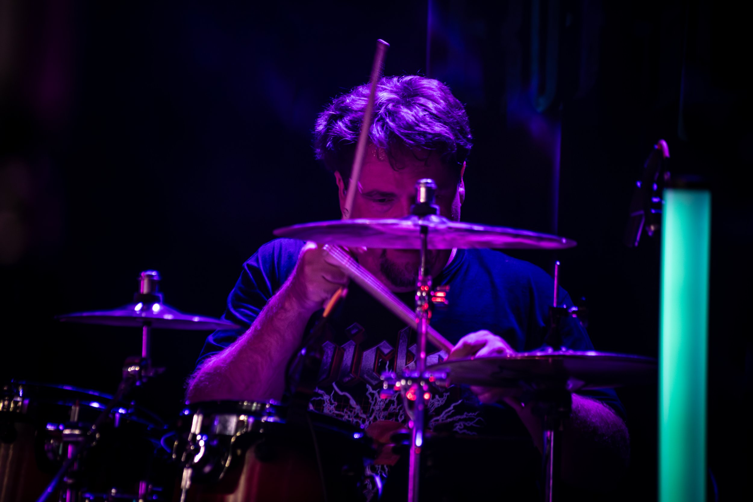 A person with dark hair playing drums in a dark setting with purple and blue lighting, with a glowing green vertical light on the right side.