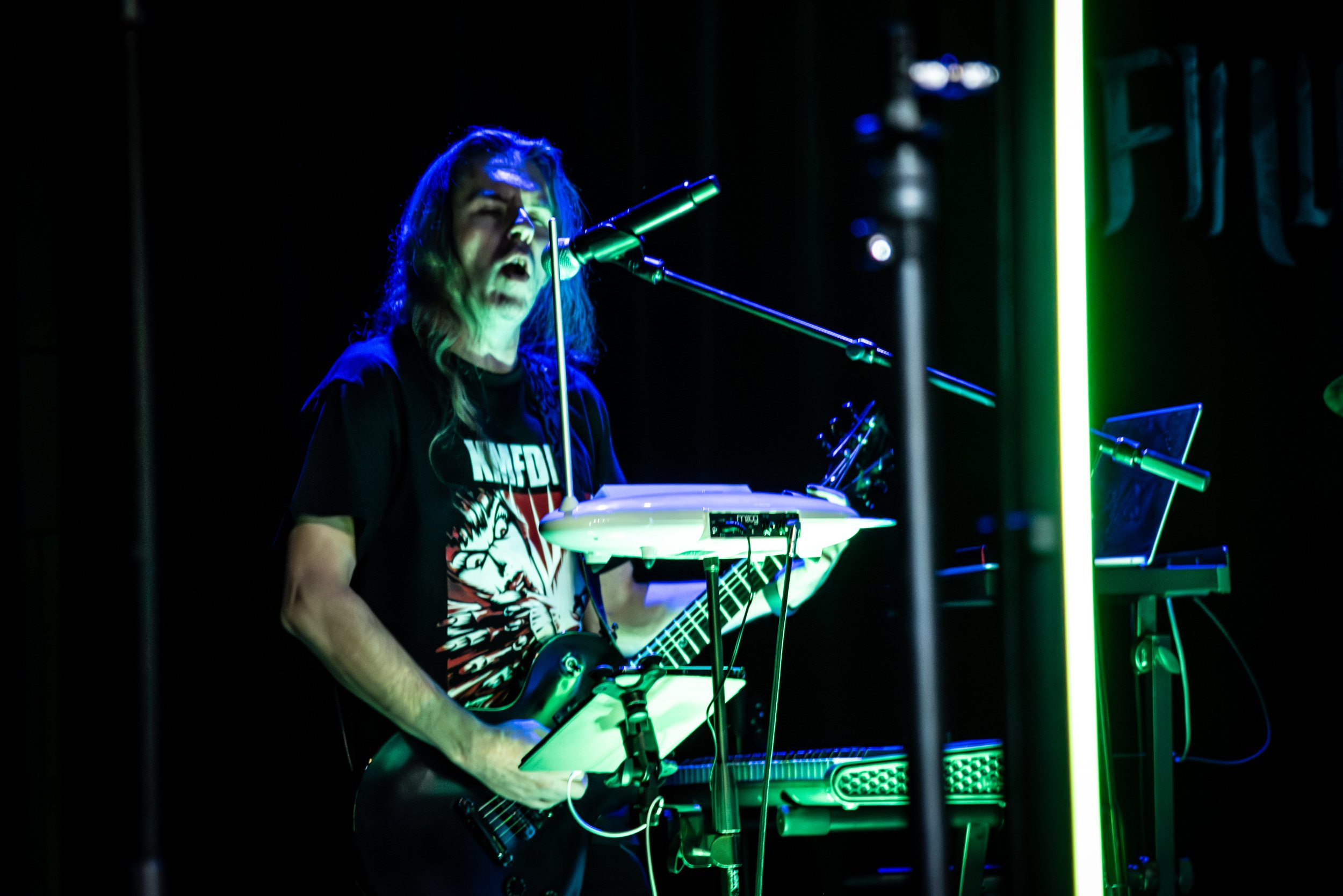 A person performing music on a stage with neon green and blue lighting, playing an electric guitar and singing into a microphone.