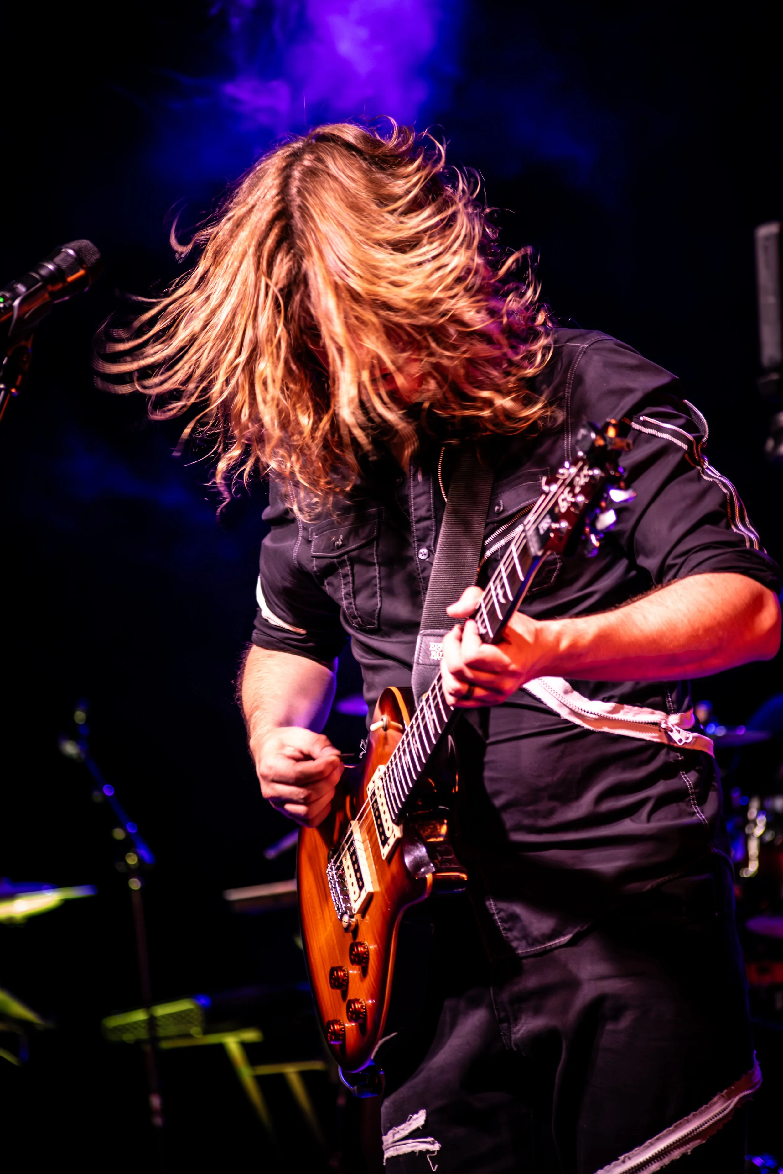 Musician with long, wavy hair playing an electric guitar on stage with purple and blue lighting.