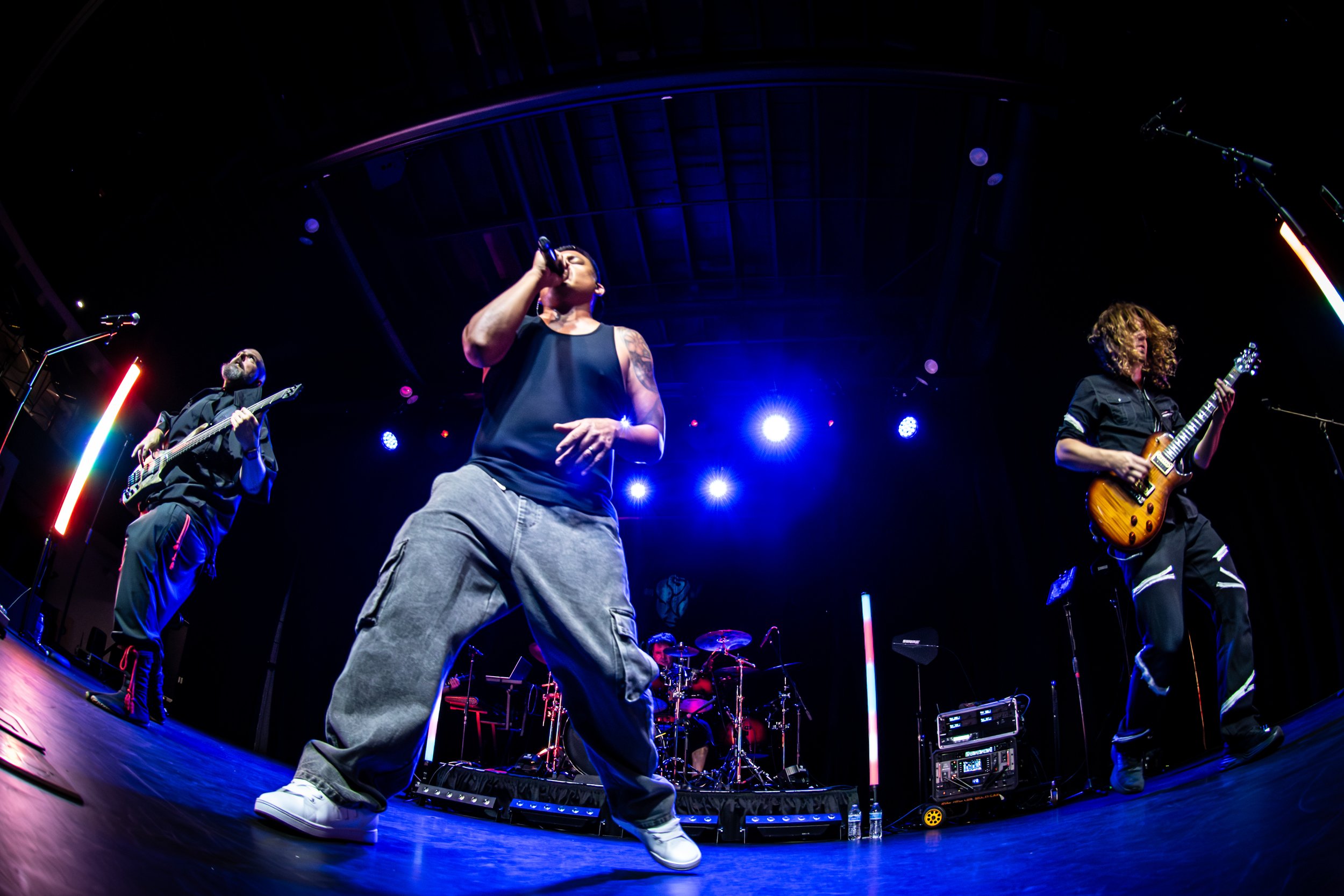 Male singer with tattoos wearing a black tank top and baggy pants singing into a microphone while performing on stage with guitarists and colorful stage lights.