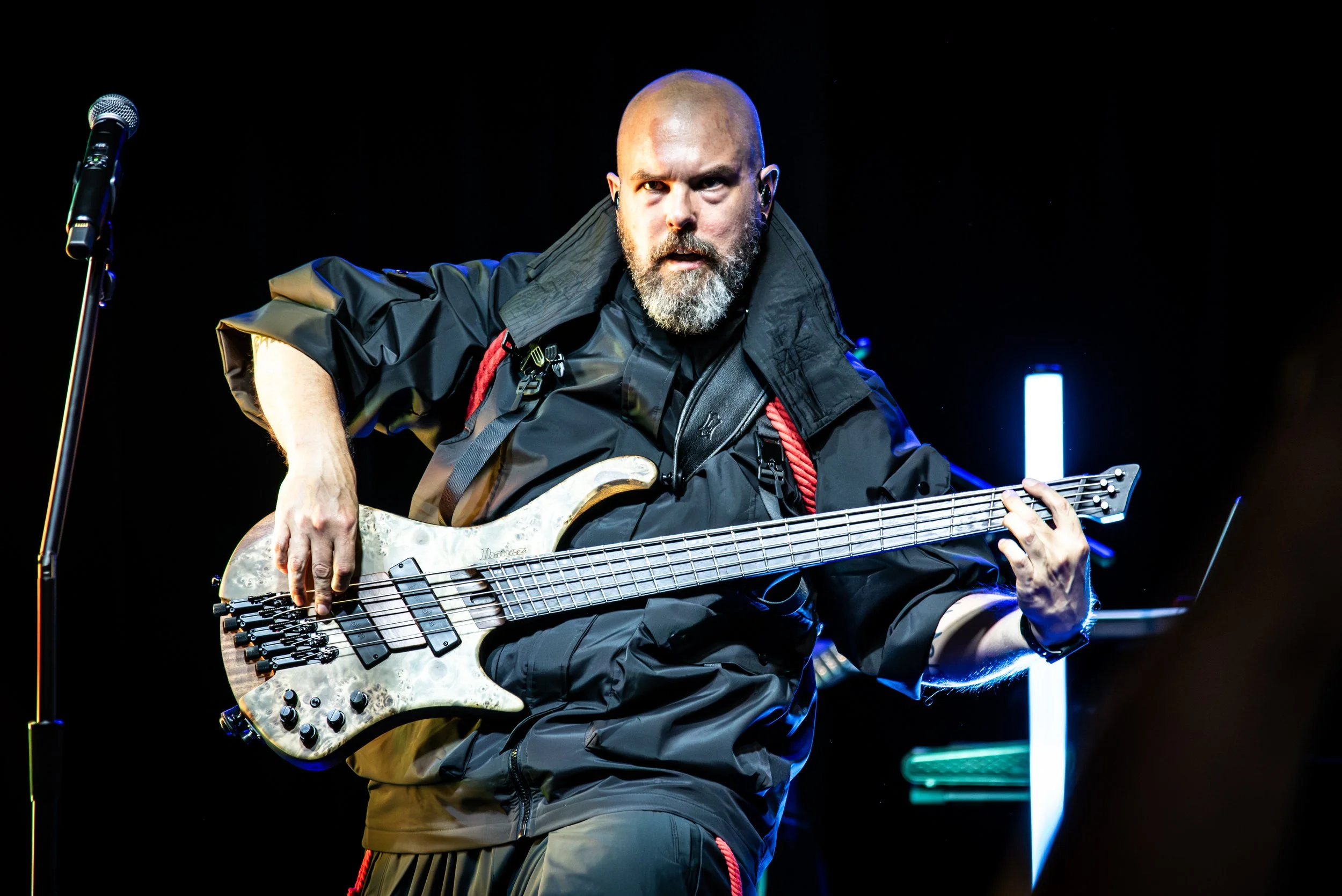 A bald man with a beard playing an electric bass guitar on stage with a dark background and faint colorful lights.