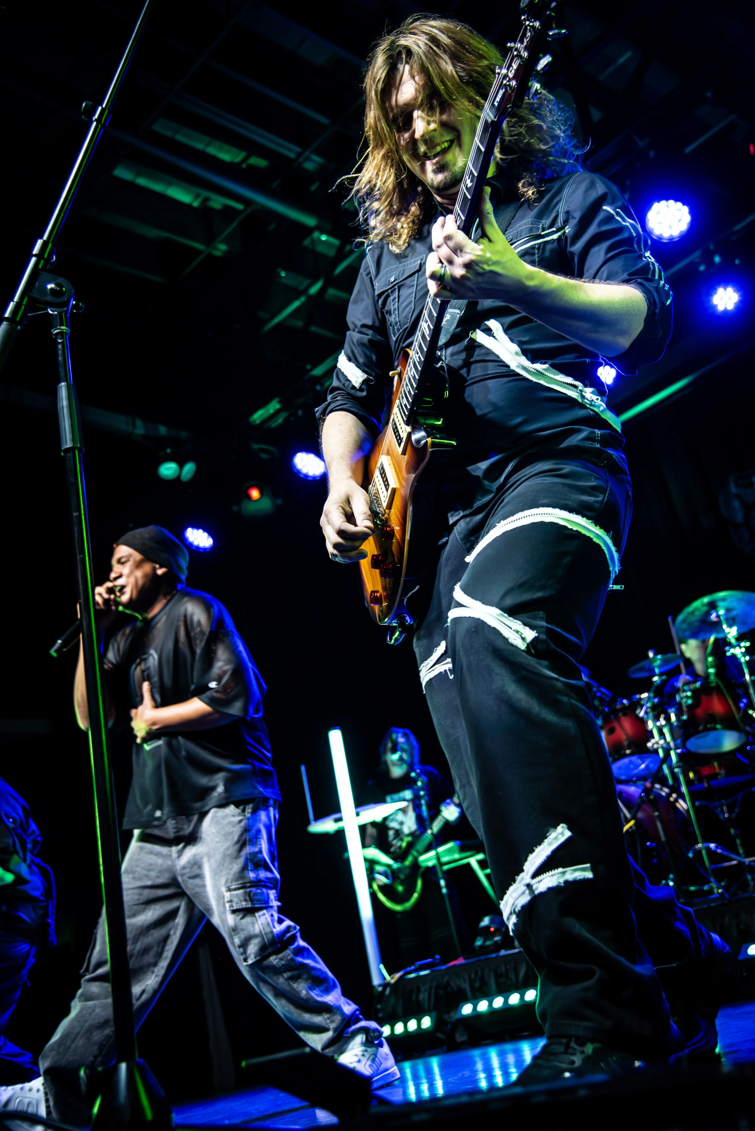 Musicians performing live on stage with neon lights, one playing an electric guitar and the other singing into a microphone.
