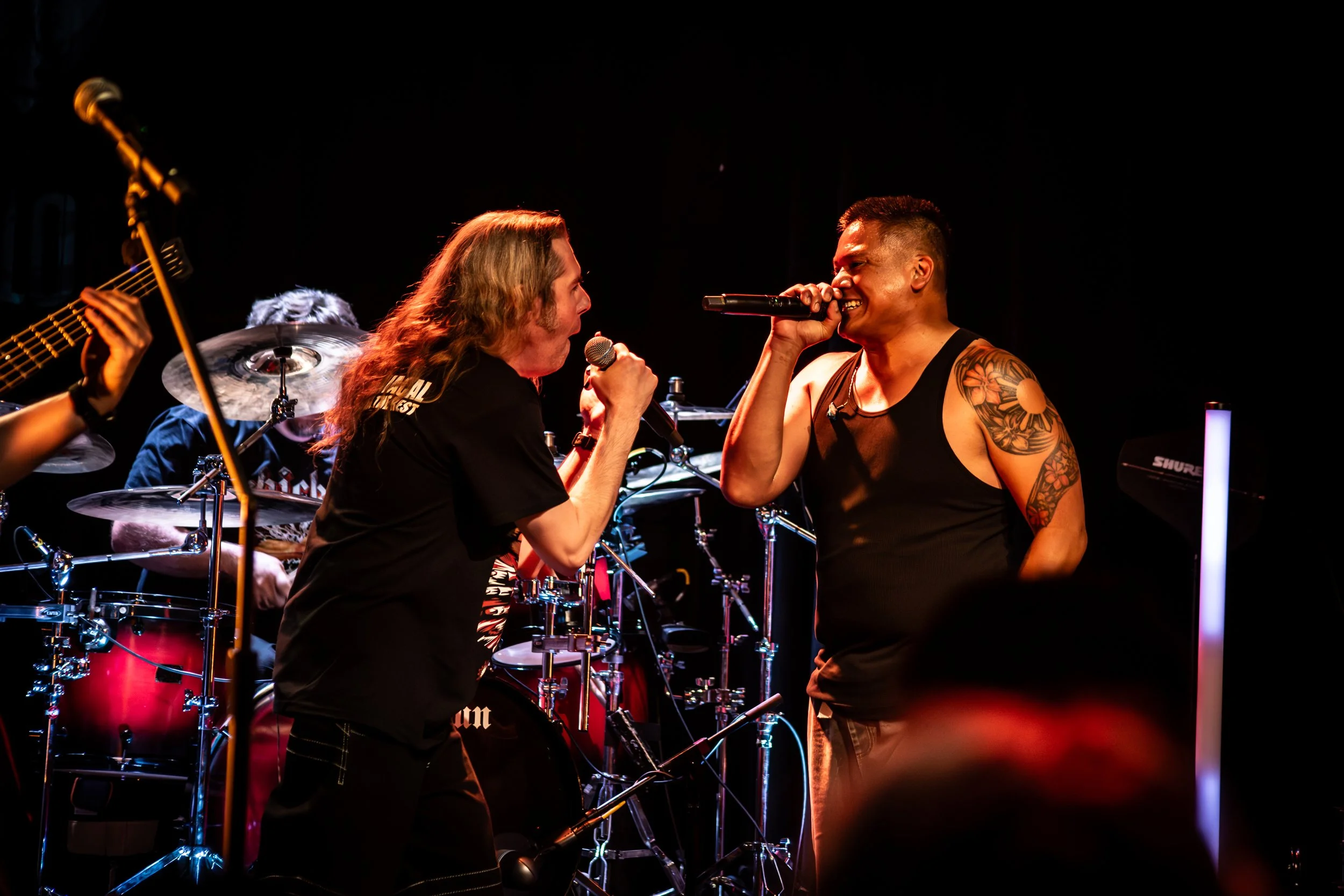 Two male singers perform on stage with a band, singing into microphones and facing each other, with a drum set and guitarist partially visible in the background.